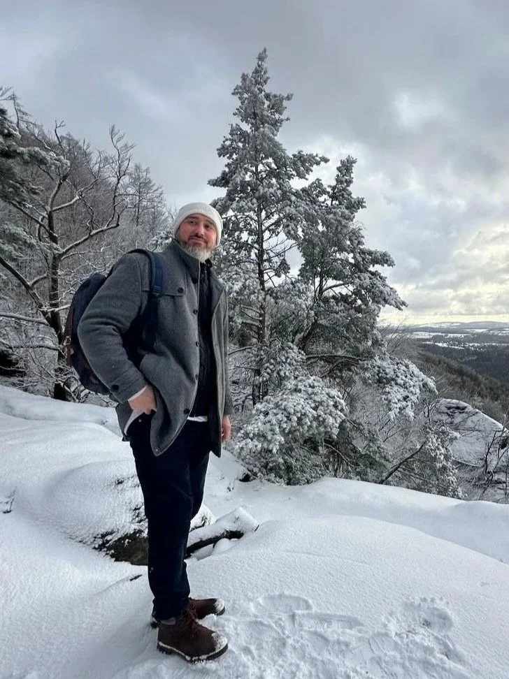 The author Yves Eugene Hensel with a gray coat, white beanie, and backpack standing in snow-covered landscape with trees and a cloudy sky.