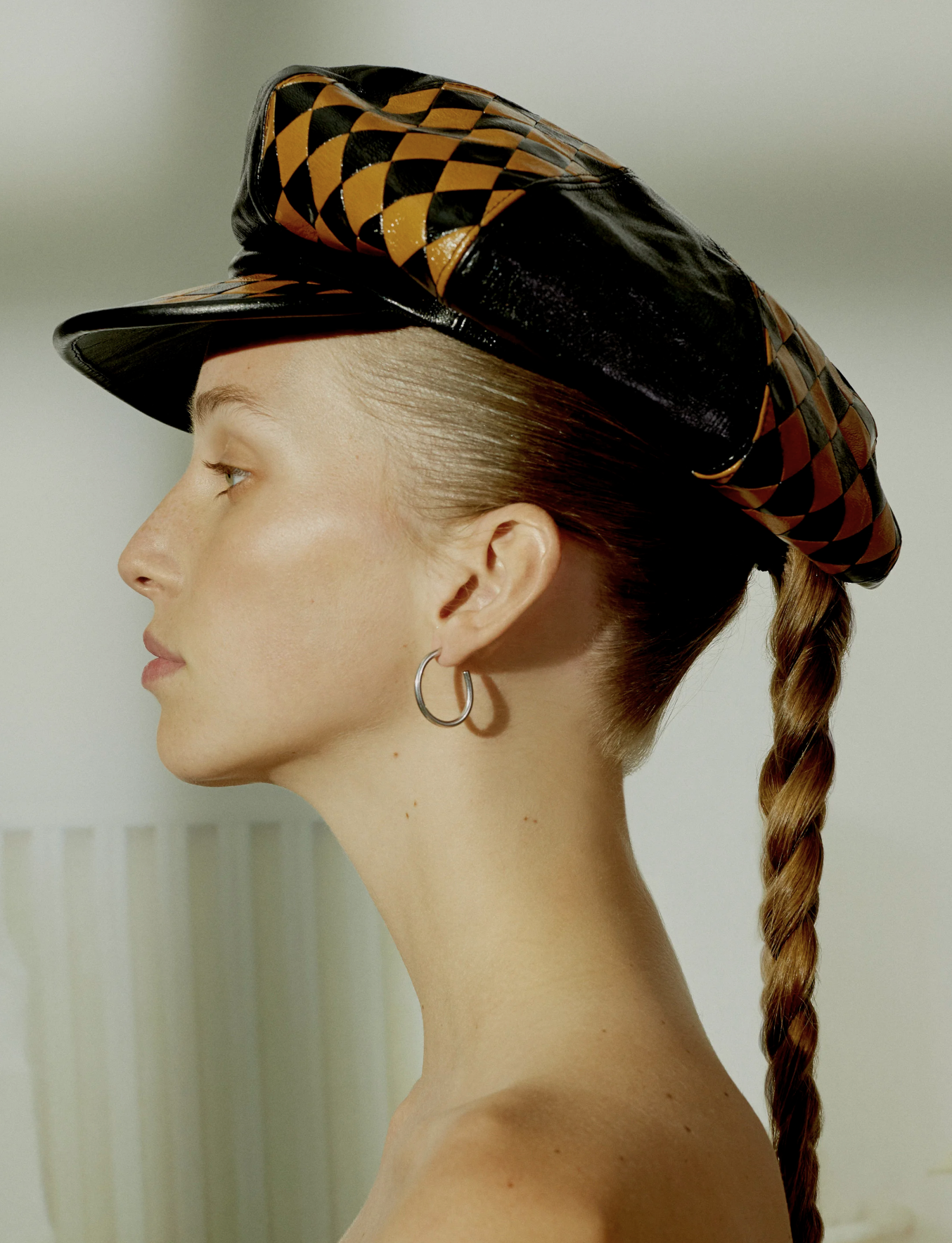 Side profile of a young woman with blonde hair in a braid, wearing a geometric patterned black and yellow leather beret and a silver hoop earring.