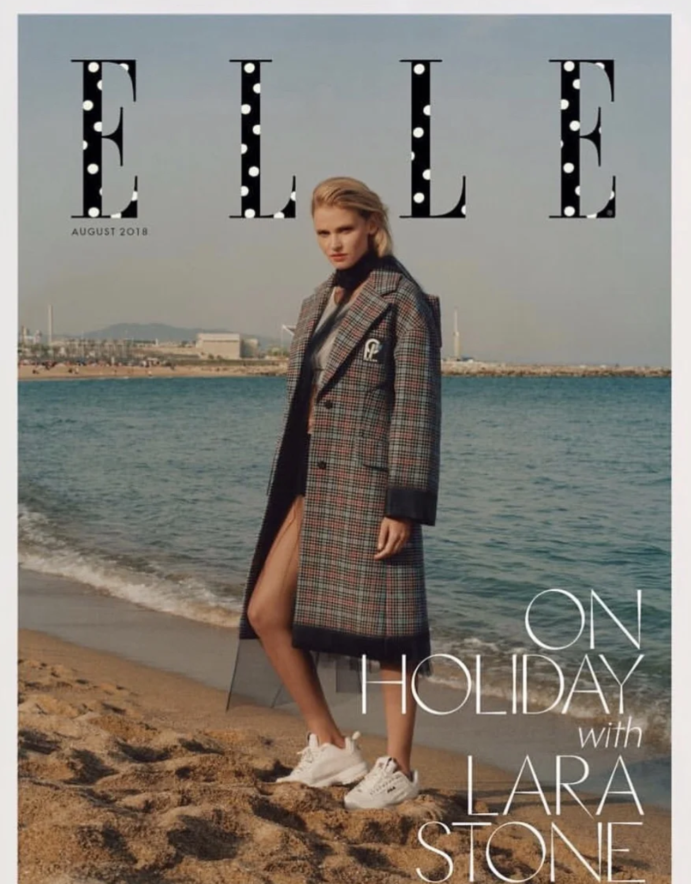 Magazine cover titled "EllE on Holiday" featuring a woman standing on a beach near the ocean, wearing a plaid coat, shorts, and sneakers, with a distant industrial landscape in the background.