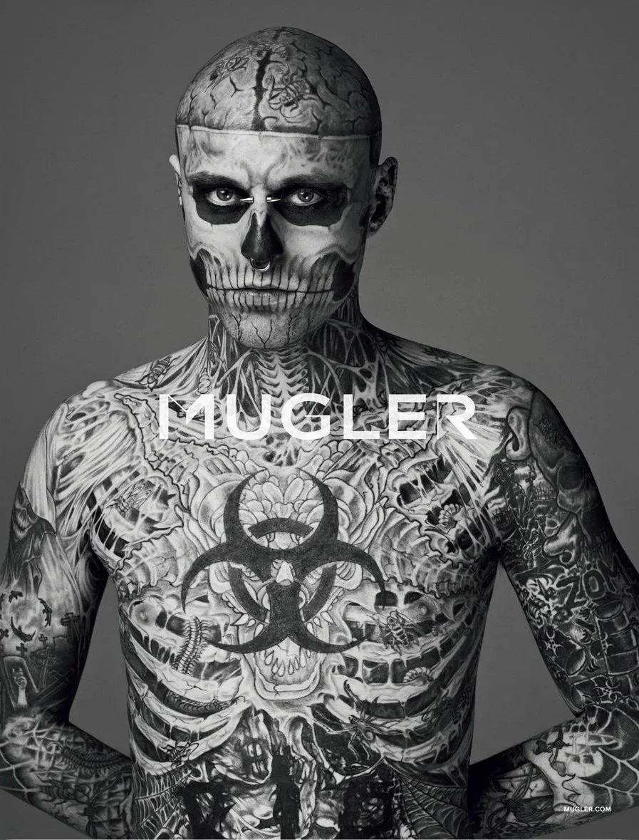 A tattooed man with a skull makeup face, bald head, and piercing, posing against a gray background. The man is covered in tattoos including skulls, biohazard symbol, and various designs.