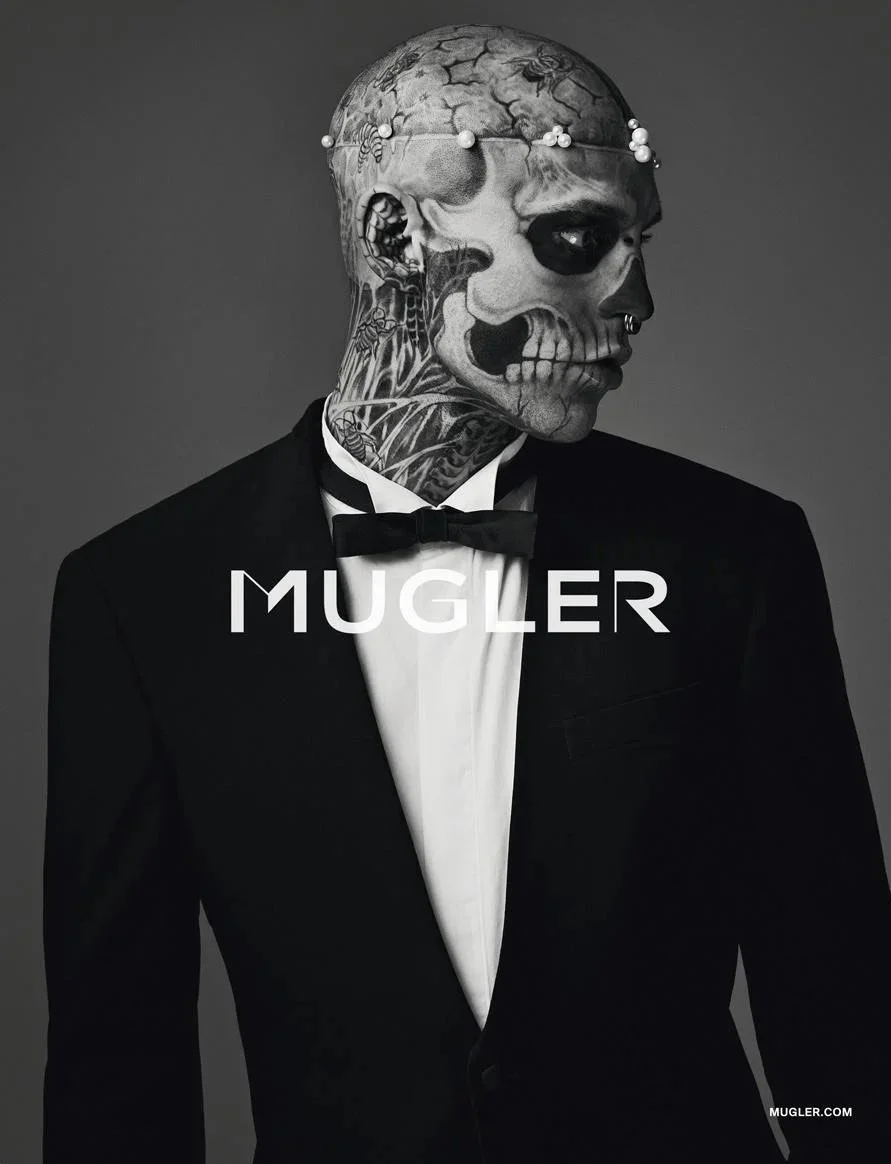 A man with tattoos and a piercings wearing a tuxedo and bow tie, with a skull face paint or mask, and a crown of pearls on his head, standing against a dark background. The word 'MUGLER' is written across the chest.