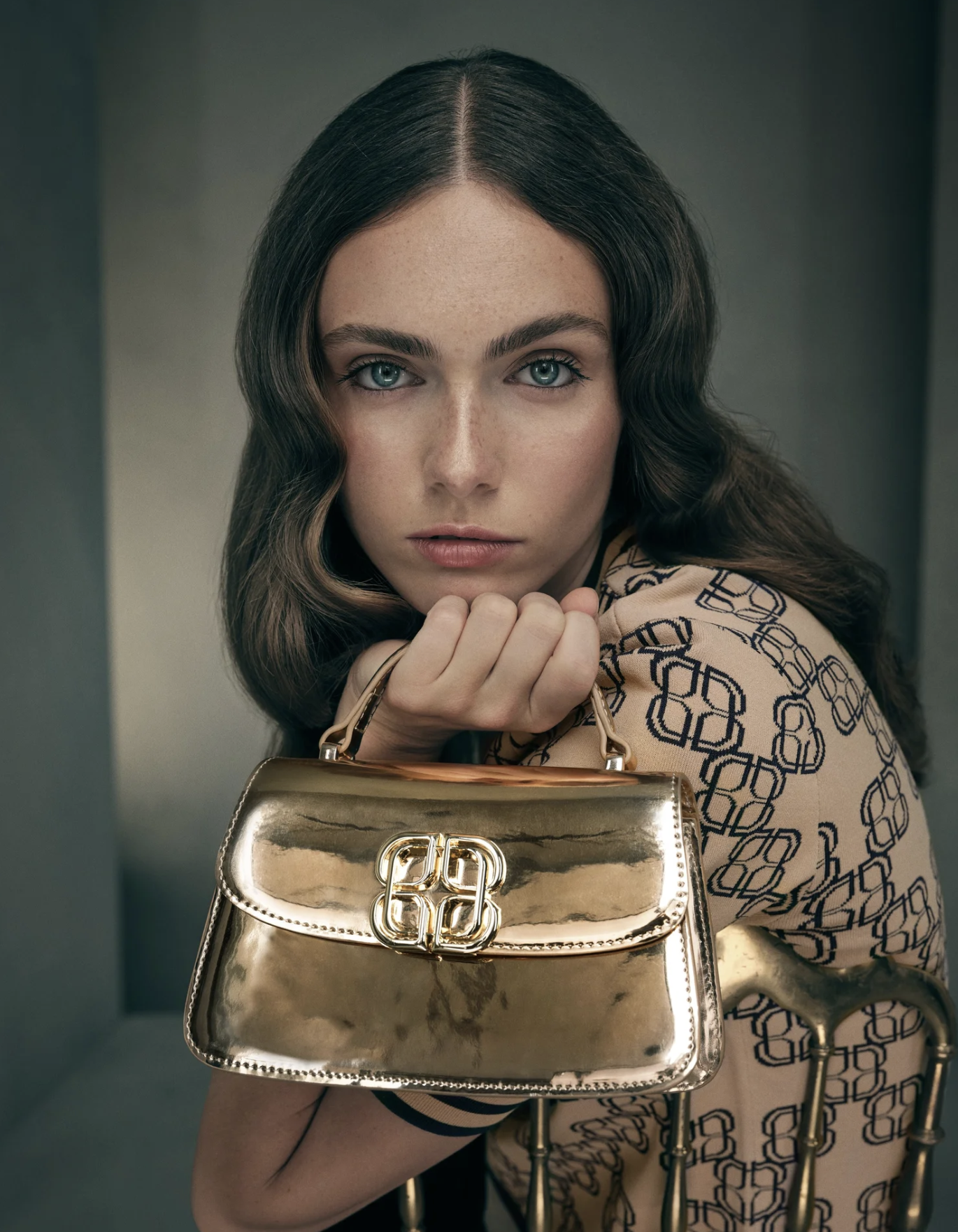 Close-up of a woman with wavy brown hair and blue eyes, holding a shiny gold designer handbag.