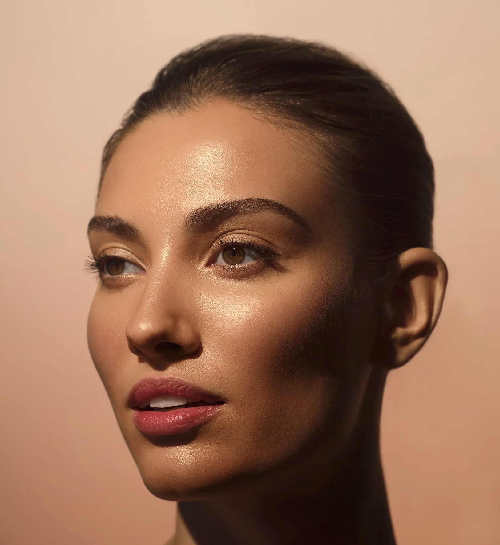 Close-up of a woman's face with smooth skin, dark hair pulled back, and minimal makeup, set against a soft beige background.