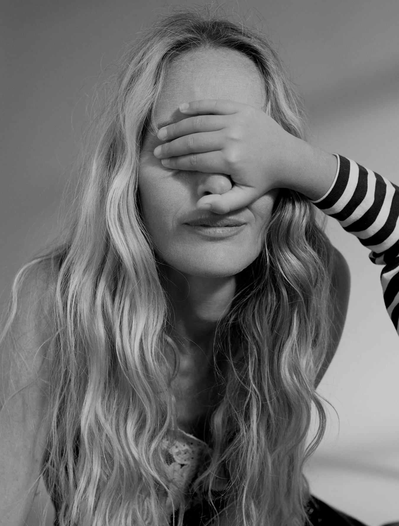 A black and white photo of a woman with long wavy hair, closing her eyes. A child's hand with long sleeves covering her face, with fingers spread, resting on the woman's forehead.