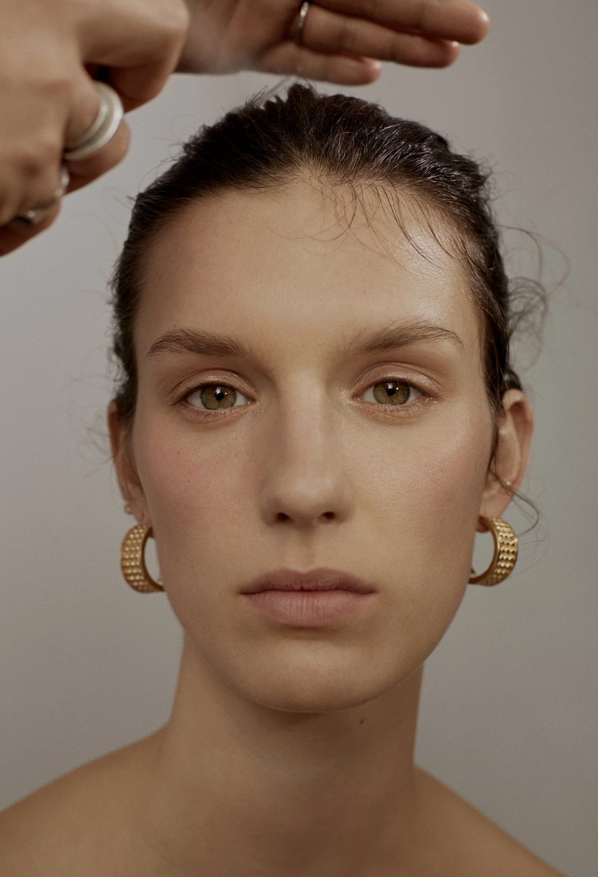 A woman with brown hair and gold hoop earrings is getting her hair styled, with her face and shoulders visible, set against a plain background.
