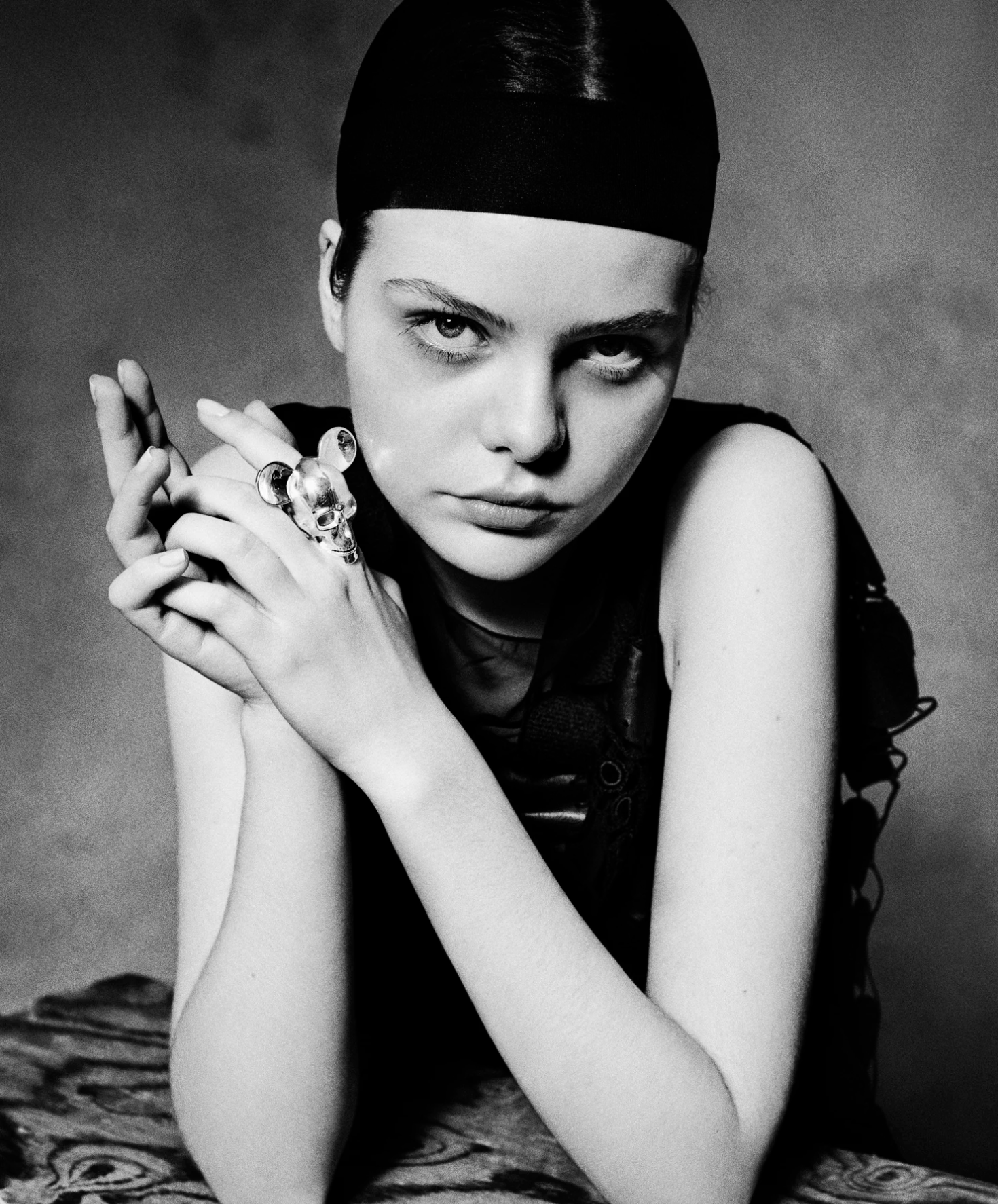 Black and white photo of a young woman with slicked-back hair wearing a head covering, looking directly at the camera, with her arms crossed and hands near her face, wearing rings and a sleeveless top, sitting on a patterned surface.