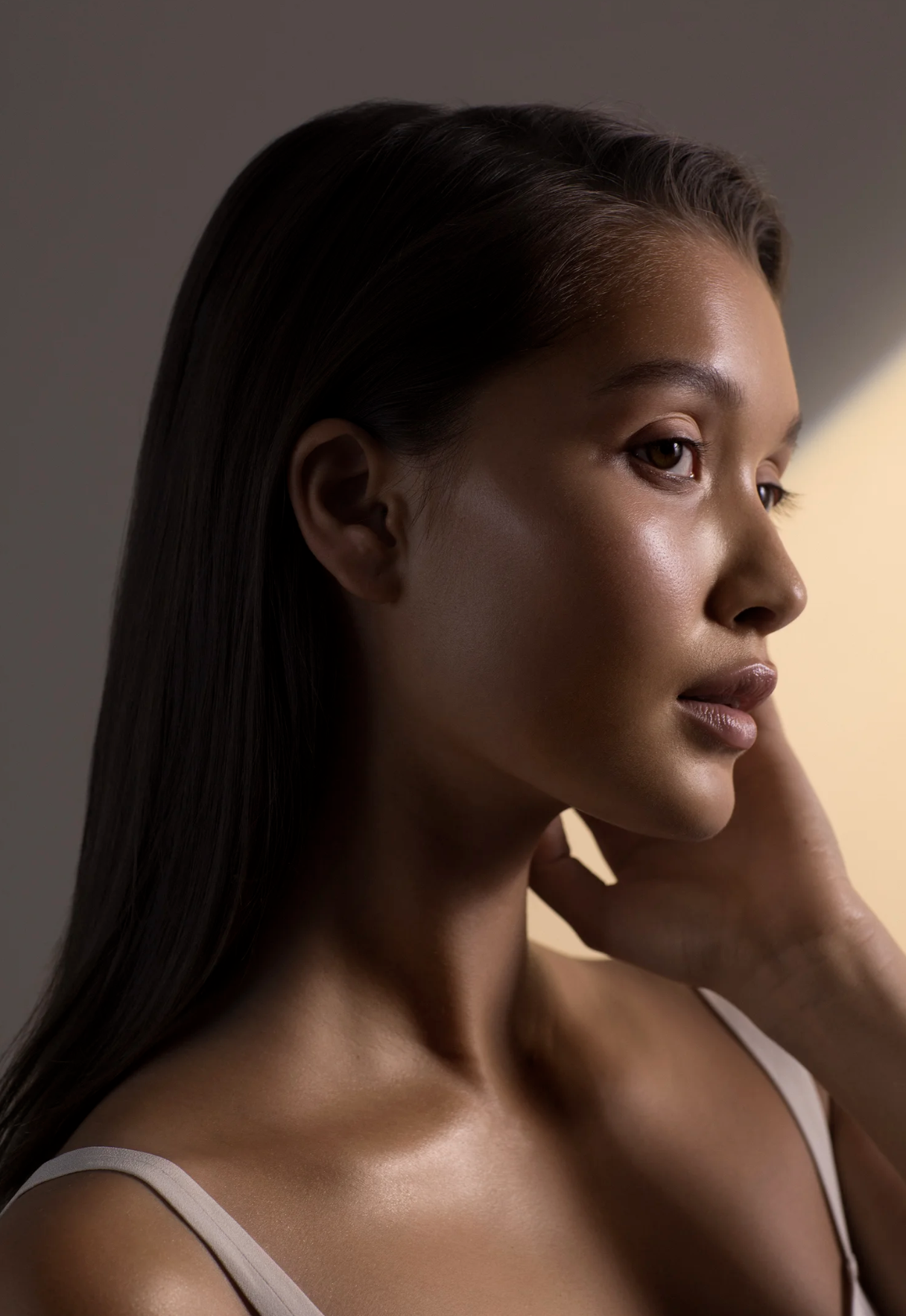 A close-up side profile of a woman with long, straight, dark hair, smooth skin, and a neutral expression, touching her neck with her left hand, wearing a light-colored sleeveless top, with soft lighting highlighting her facial features.