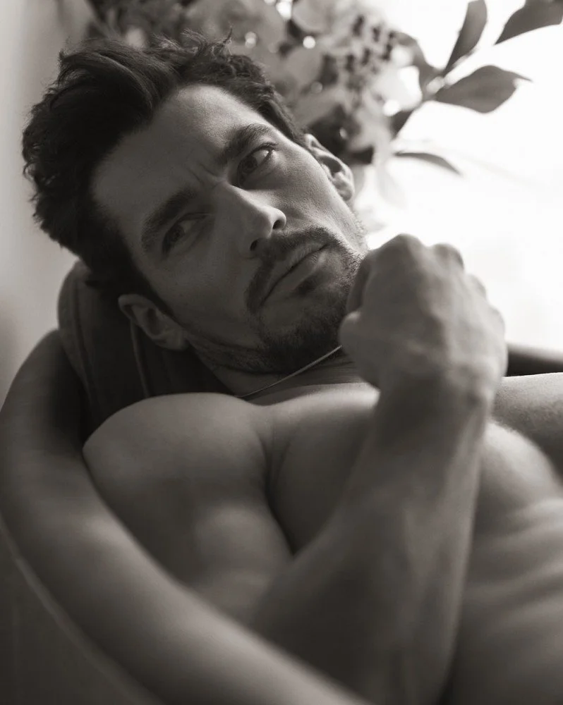A black and white portrait of a muscular man lying down, looking to the side with a contemplative expression, and his arm flexed to show his bicep.