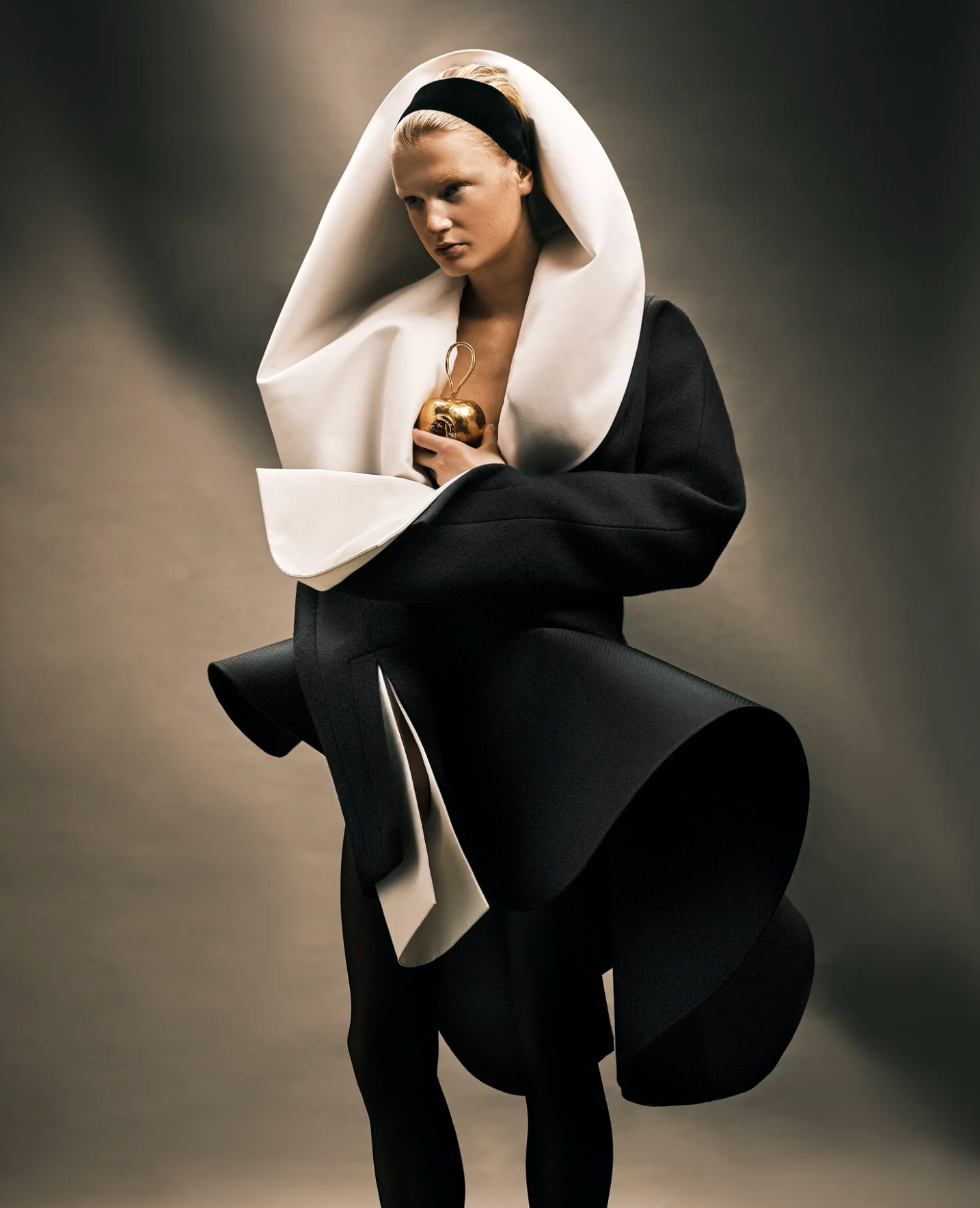 A woman with blonde hair and black headband wearing a black and white avant-garde outfit, holding a gold apple with a loop handle, against a gradient gray background.