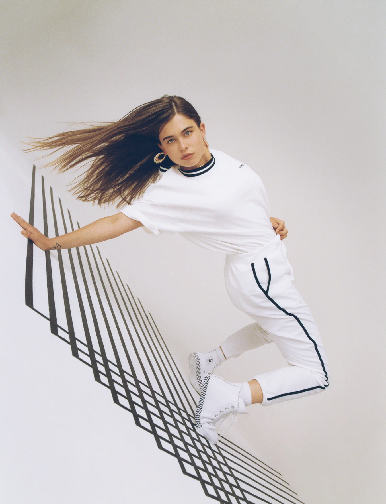 A young woman with long hair and hoop earrings poses against a white background, with shadow lines creating a geometric pattern.