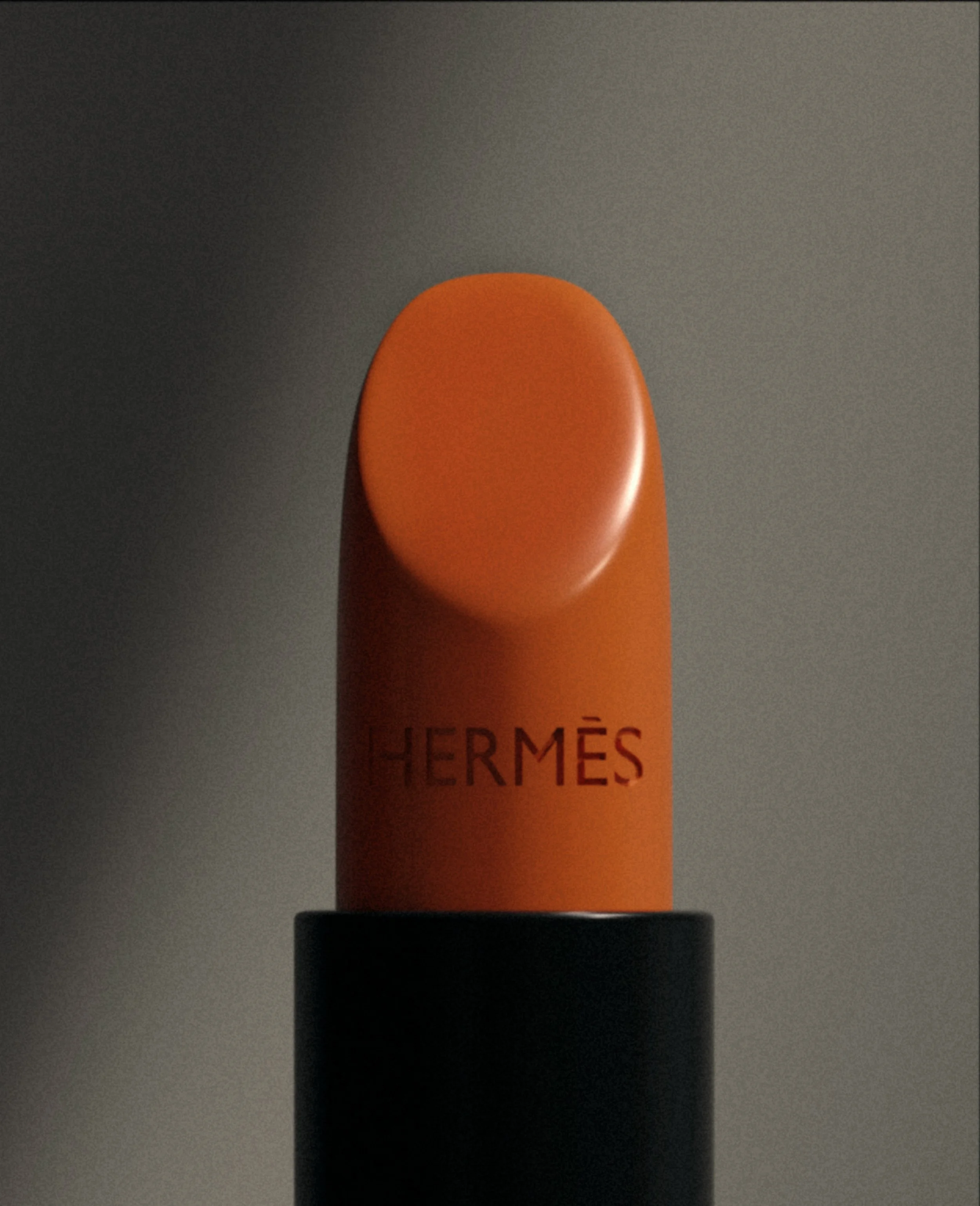 Close-up of an orange Hermès lipstick with the logo visible on the tube, placed on a dark surface against a dark background.