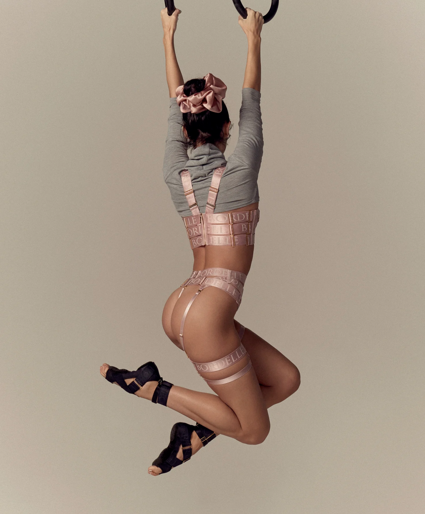 A person jumping with arms stretched upward holding onto gymnastic rings, wearing a beige suspender set with "BELLE" printed, a gray top, and black sandals, with a plain background.