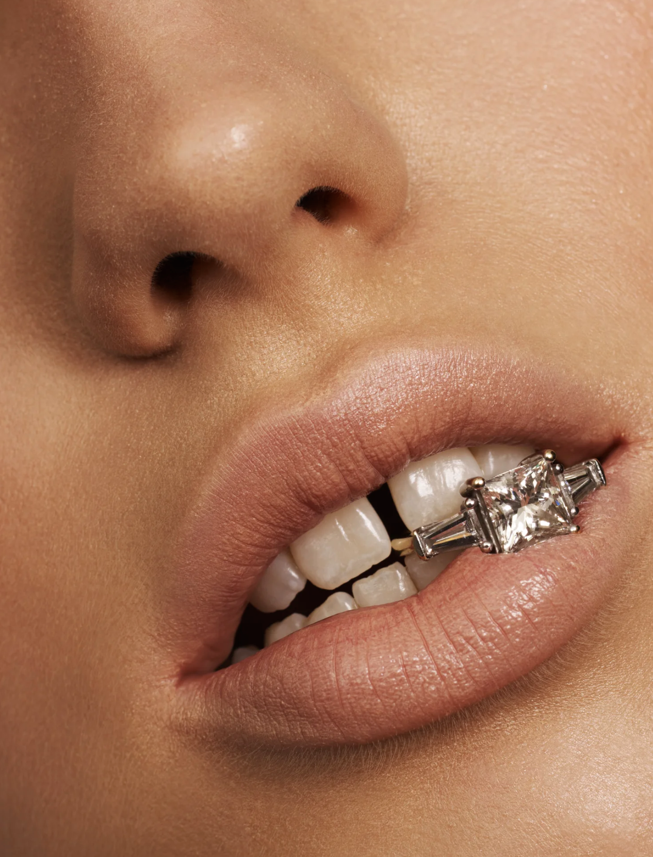 Close-up of a person's face showing their nose and lips, with a large square-cut diamond ring on the teeth between their front teeth.