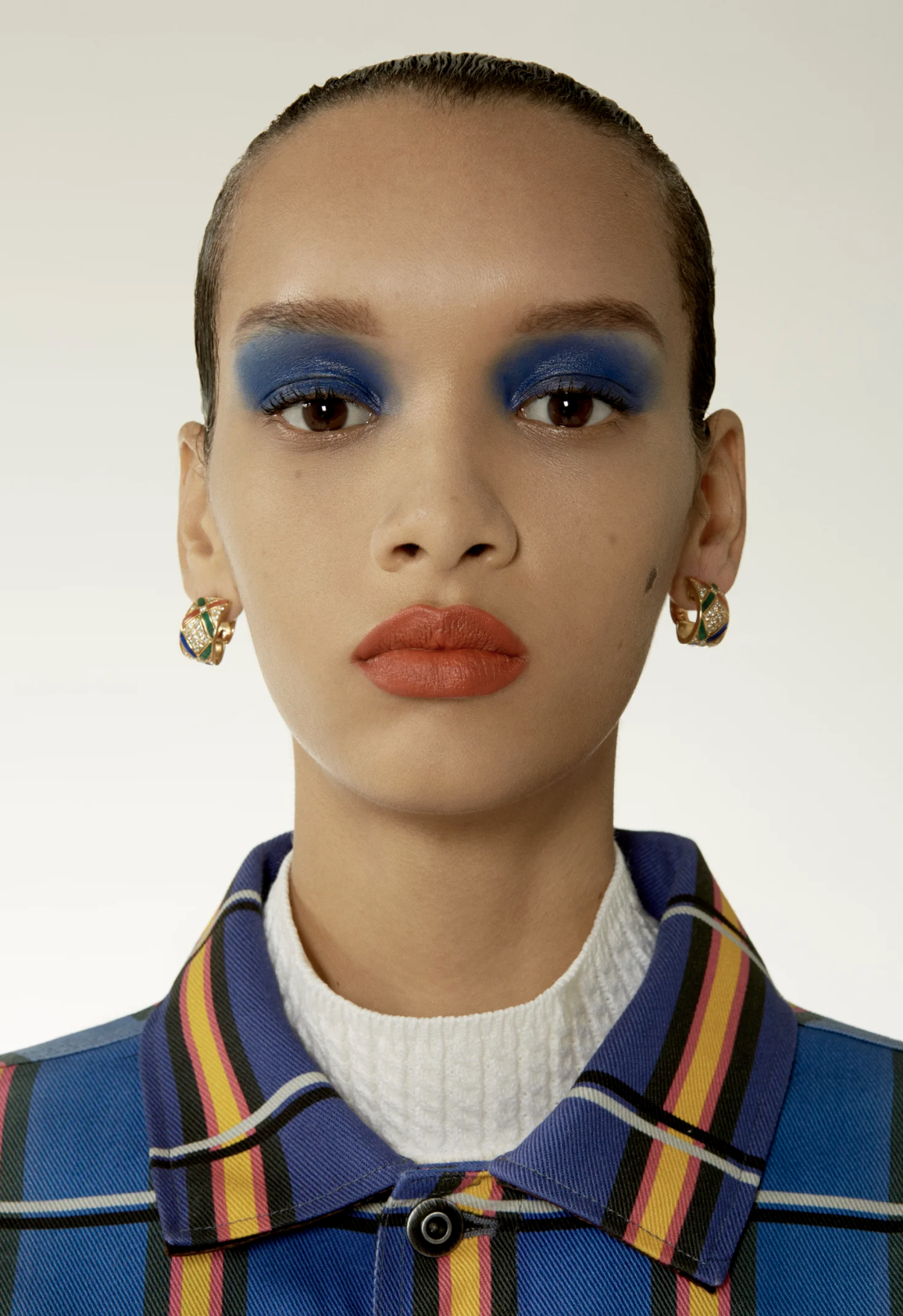 Close-up of a woman with bold blue eye makeup, red lipstick, wearing colorful earrings and a plaid jacket with a white turtleneck underneath.
