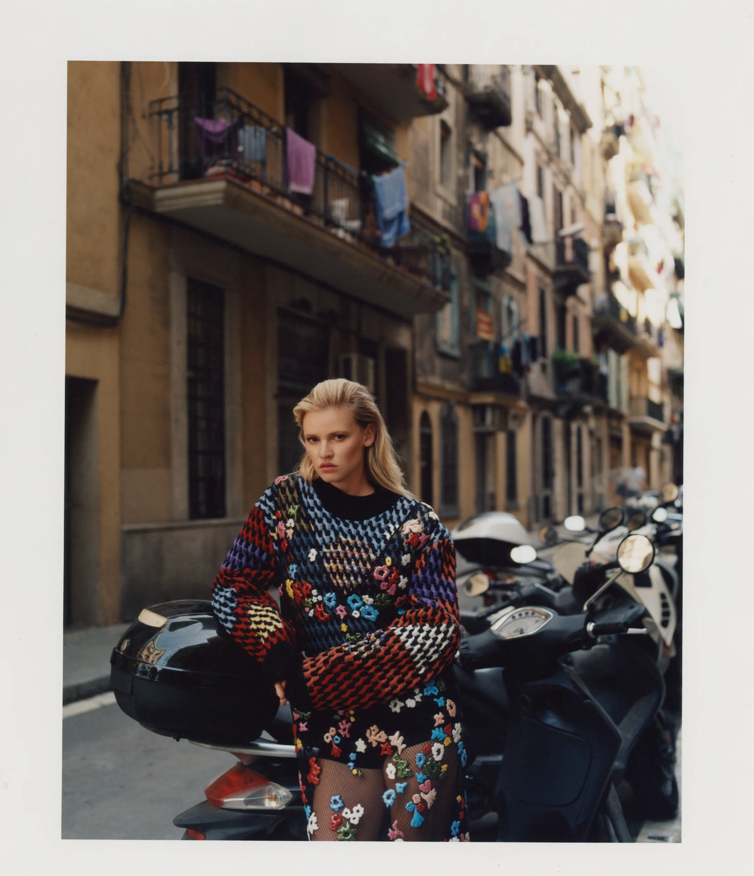 A young woman with blonde hair styled in a side part, standing next to a black scooter, wearing a colorful, embroidered dress with floral patterns and fishnet stockings, on an urban street with apartment buildings and parked scooters.