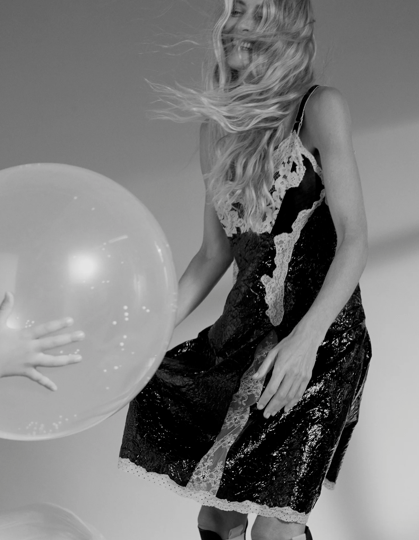 A woman with long wavy hair smiling and playing with a large, clear balloon while wearing a lace and sequin dress.