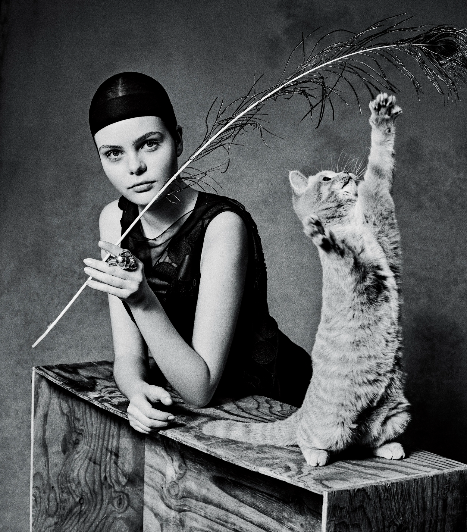A black and white photo of a woman with dark headscarf and dark dress sitting on a wooden platform holding a large feather, while a tiger cub reaches up with its paw to touch the feather.
