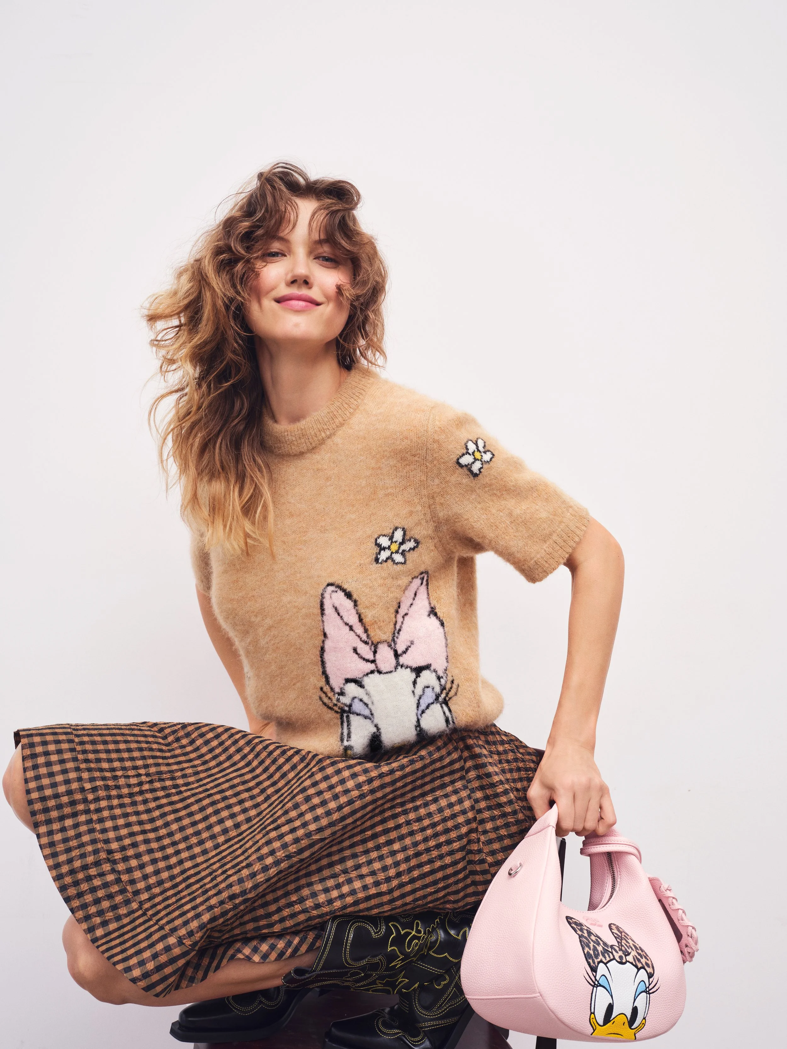 A woman with wavy brown hair in a beige sweater with Daisy Duck design, plaid skirt, black boots, holding a pink handbag with Daisy Duck print on it, against a plain white background.