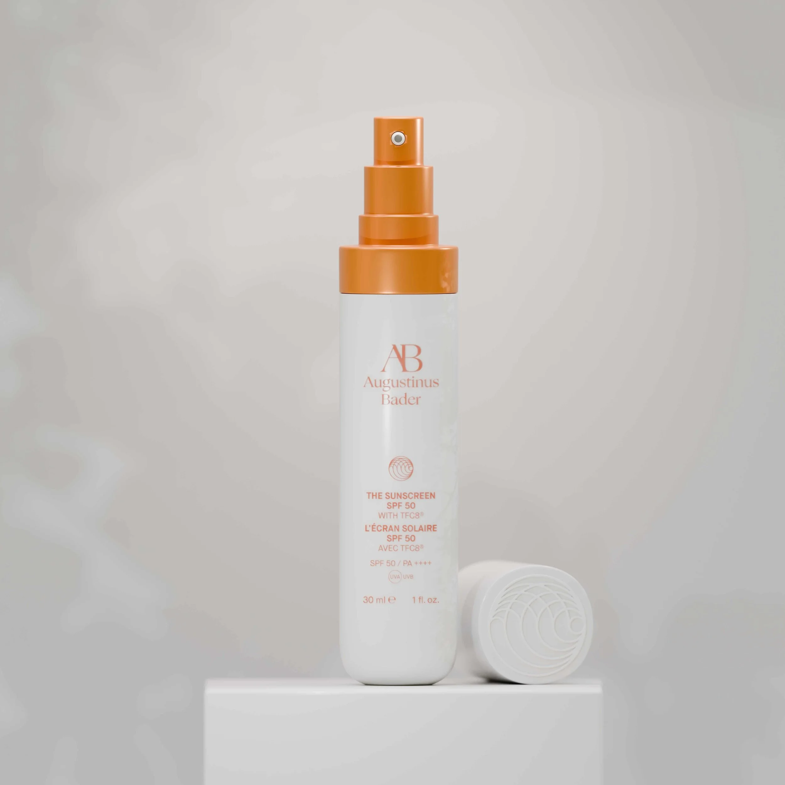 A white bottle of sunscreen with orange writing and an orange cap, placed on a white surface. The bottle is from the brand Augustinus Bader and the label reads 'The Sunscreen SPF 50' with some additional product information. A white cap is lying next