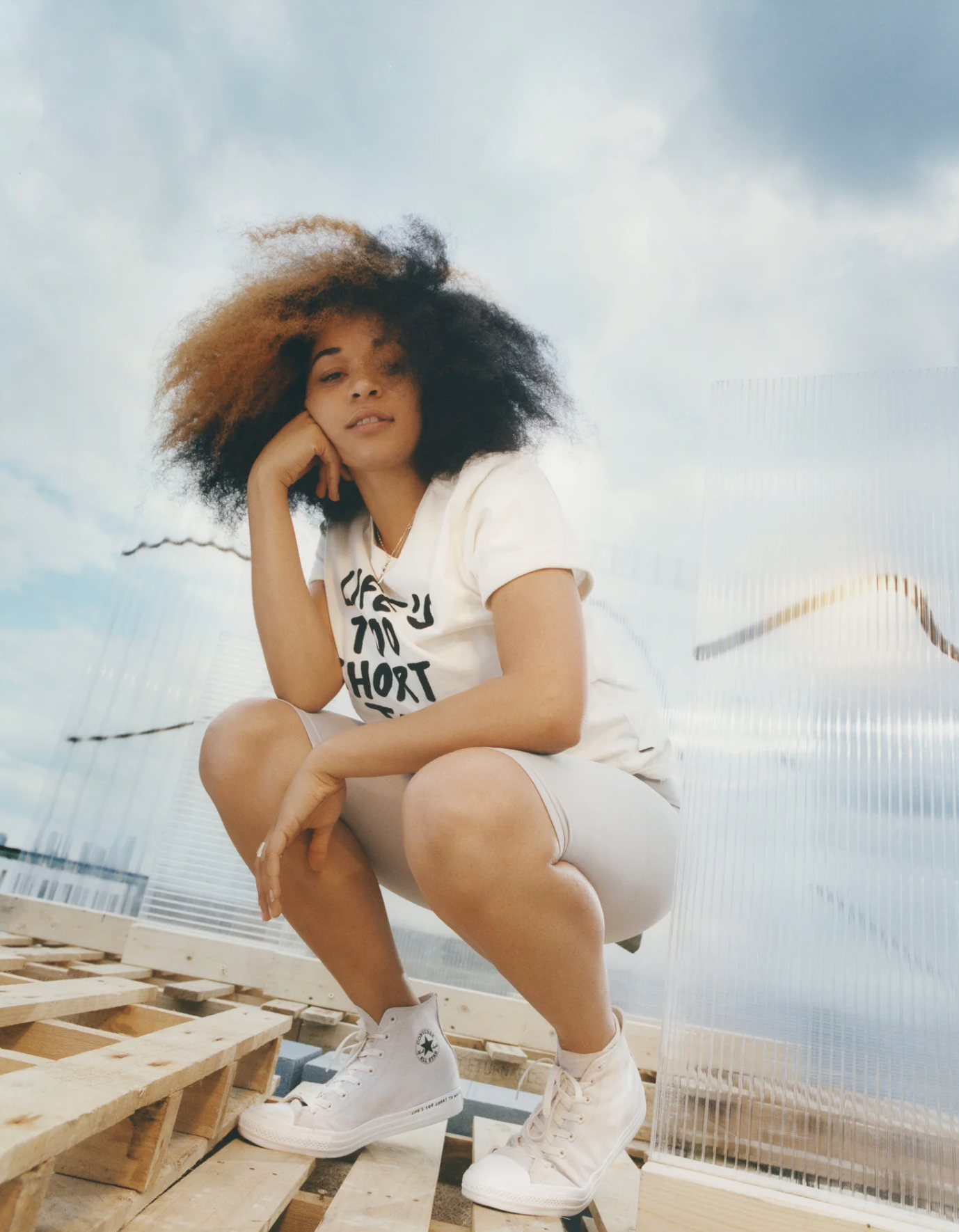 Young woman with curly hair squatting on wooden pallets under a cloudy sky, wearing white Converse sneakers, shorts, and a T-shirt.