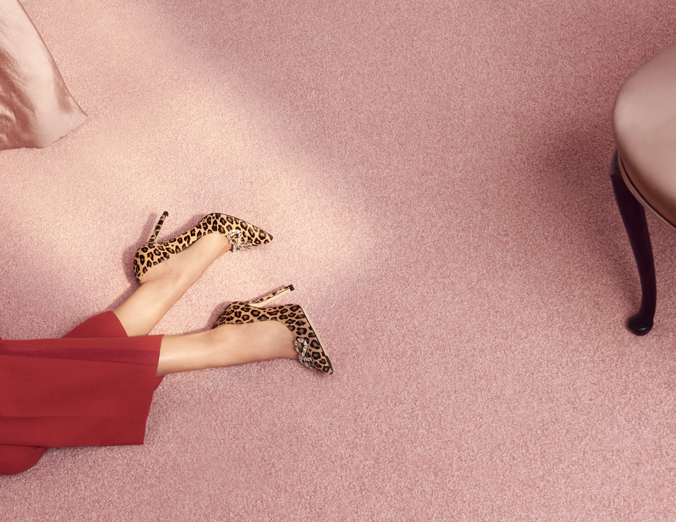 A woman wearing red dress and leopard print high heels standing on pink textured carpet, with part of a white pillow visible on the left side and a purple chair on the right side.