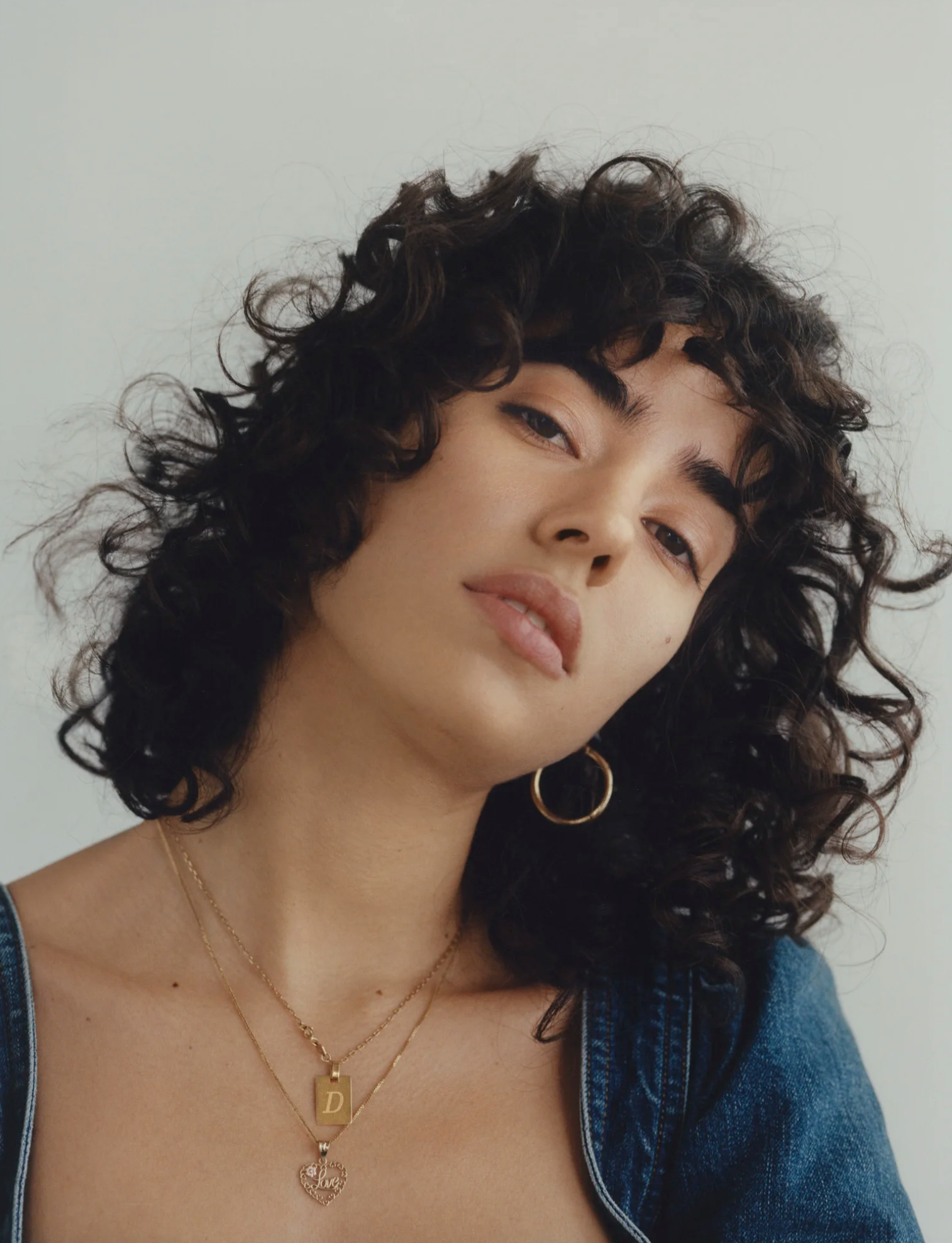 Close-up of a woman with curly dark hair, wearing layered gold necklaces and gold earrings, dressed in a denim jacket.