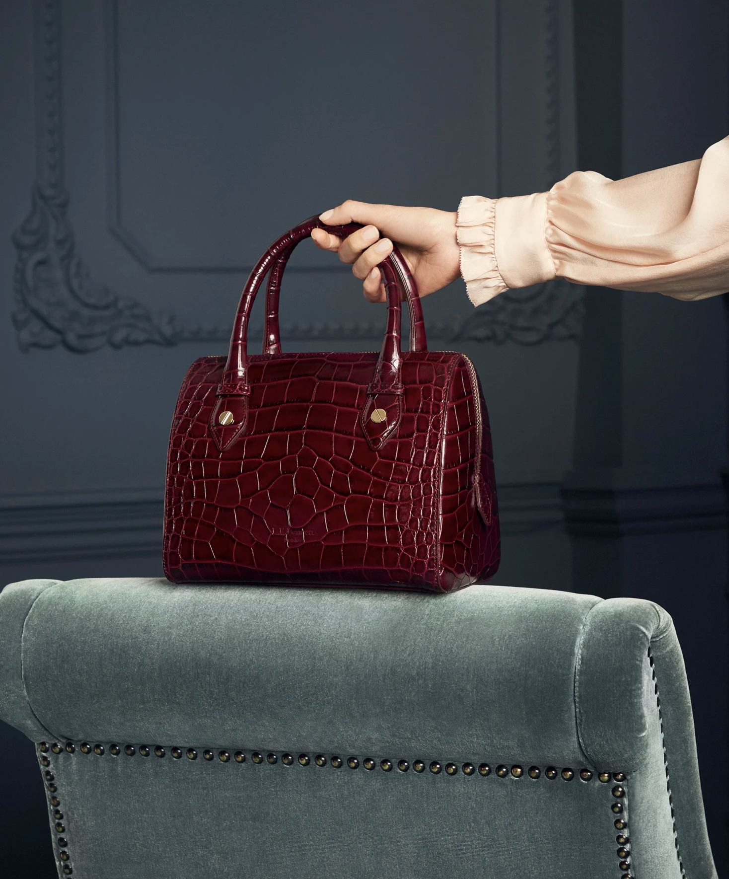 A red crocodile-embossed handbag resting on a green velvet armrest of a sofa. A person's hand, wearing a beige long-sleeve shirt, is holding the handbag's handle. The background includes decorative wall molding.