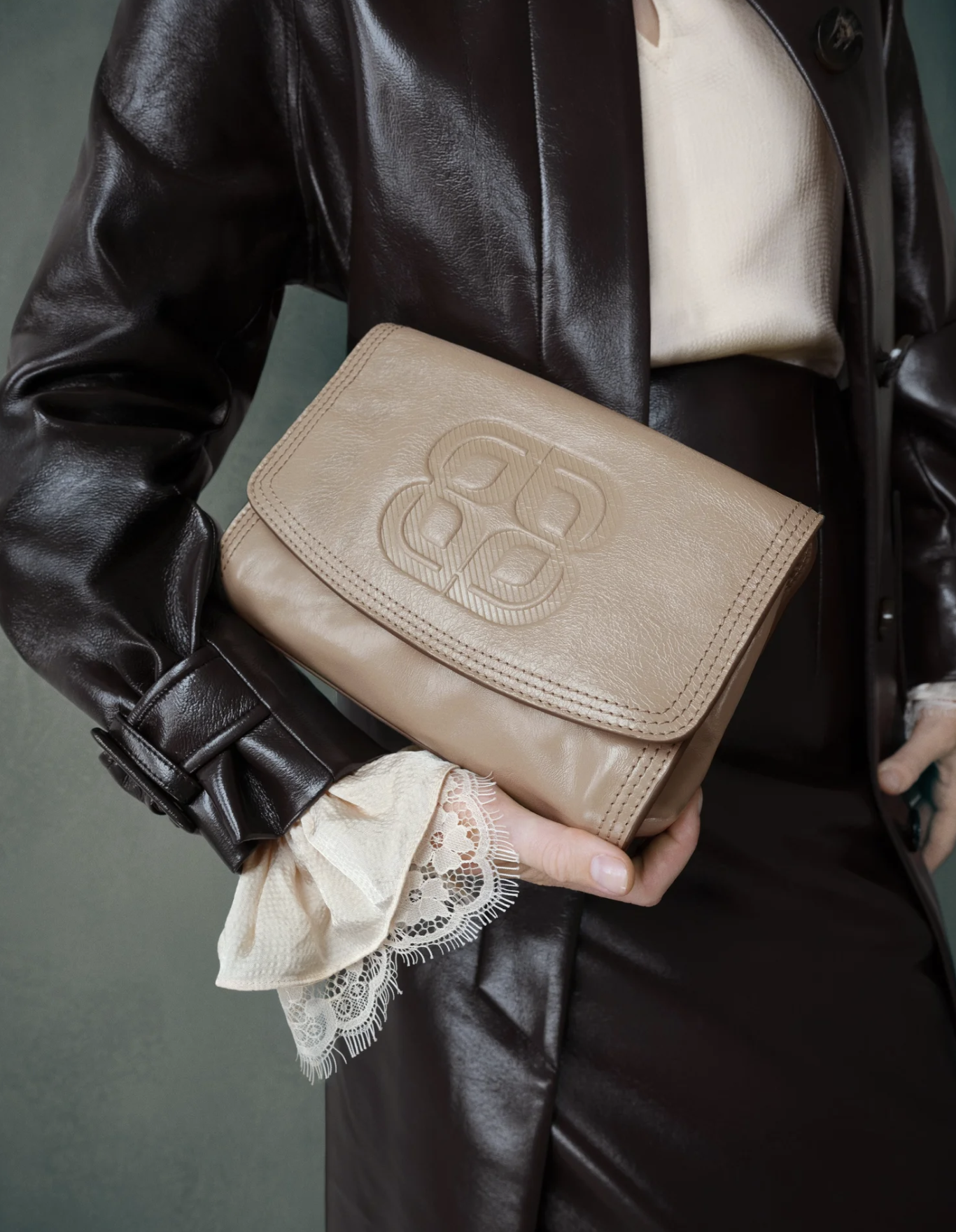 Person wearing a black leather jacket and cream-colored sweater holding a beige designer clutch with an embossed logo, with another person wearing a lace cuff visible.