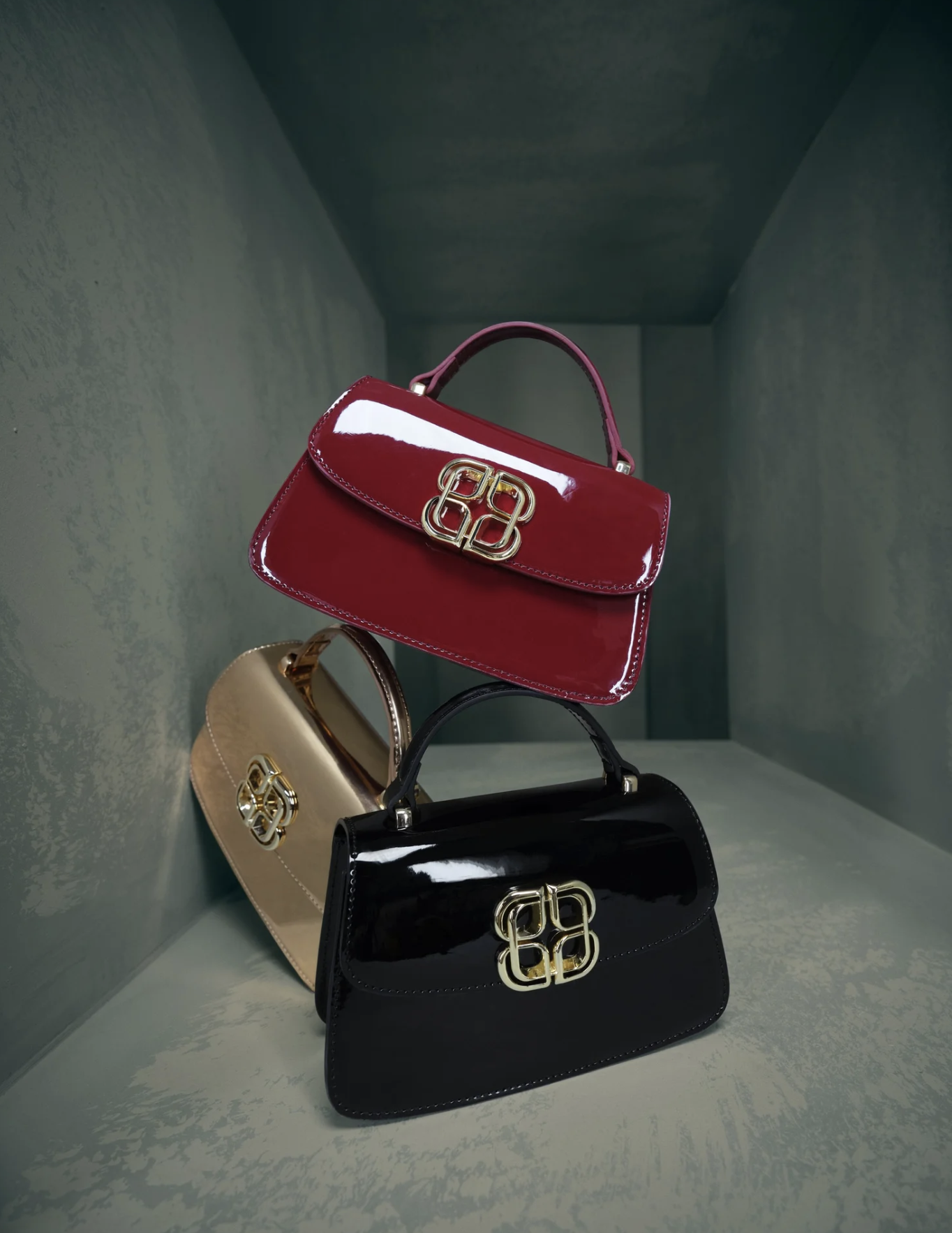 Display of four luxury handbags in black, beige, pink, and black, each with a gold logo on the front, arranged on a green shelf.
