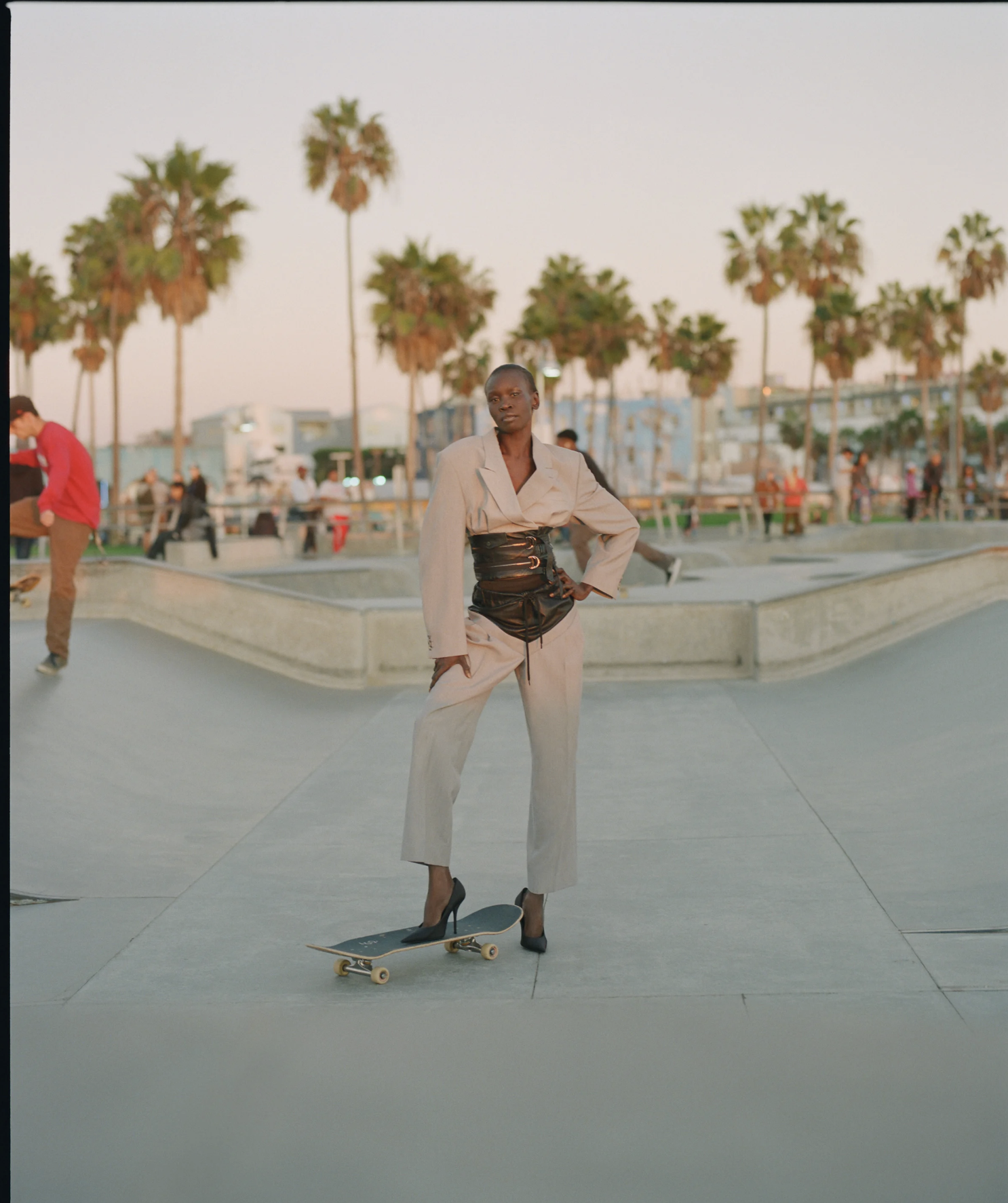 A woman in beige pants and blazer, wearing high heels, stands on a skateboard at a skate park during sunset, with palm trees and other people in the background.