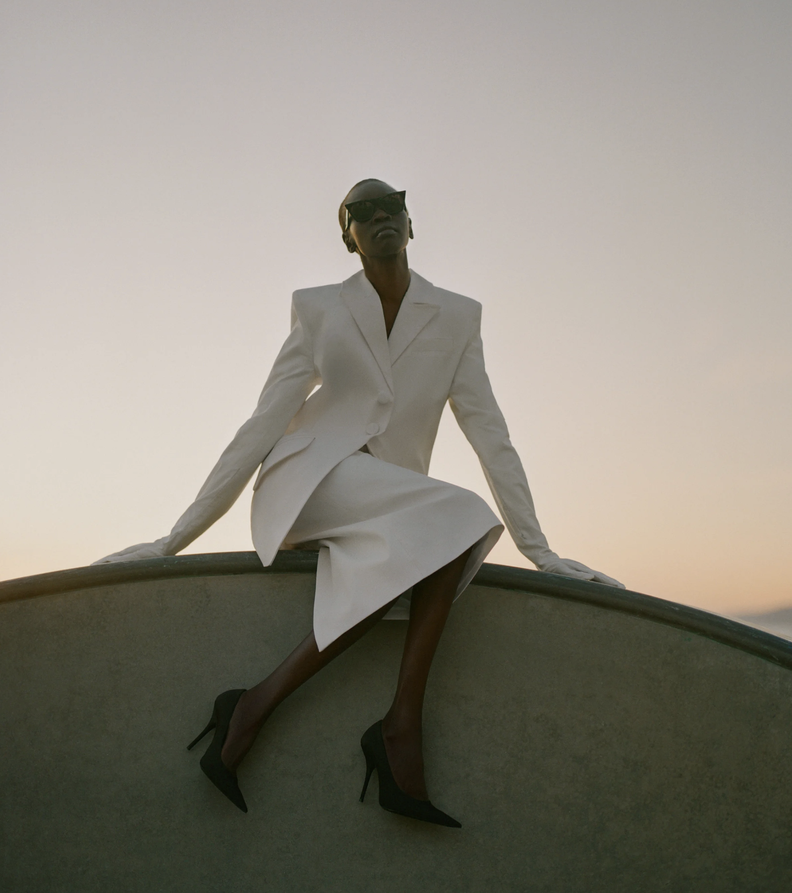 A woman wearing a white suit, black heels, and sunglasses, sitting on a curved surface outdoors at sunset or sunrise.