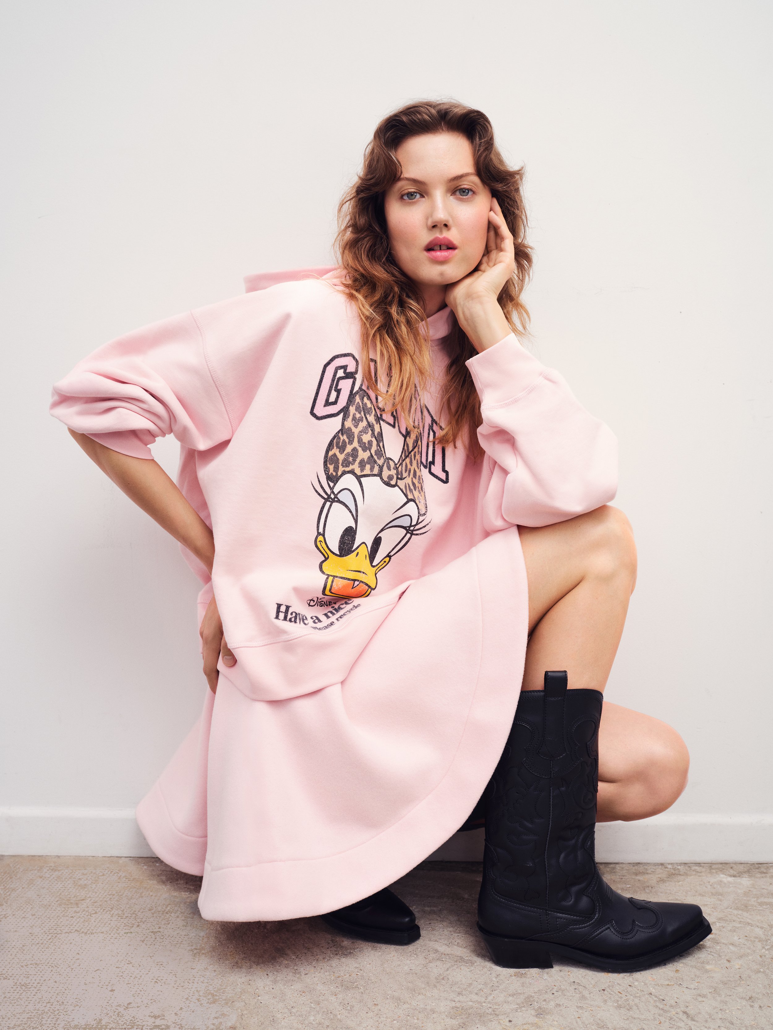 A woman with light skin, wavy brown hair, and blue eyes is squatting against a white wall. She is wearing a pink oversized hoodie with a graphic of Daisy Duck and leopard print, black thigh-high cowboy boots, and black knee-high socks with a pattern.