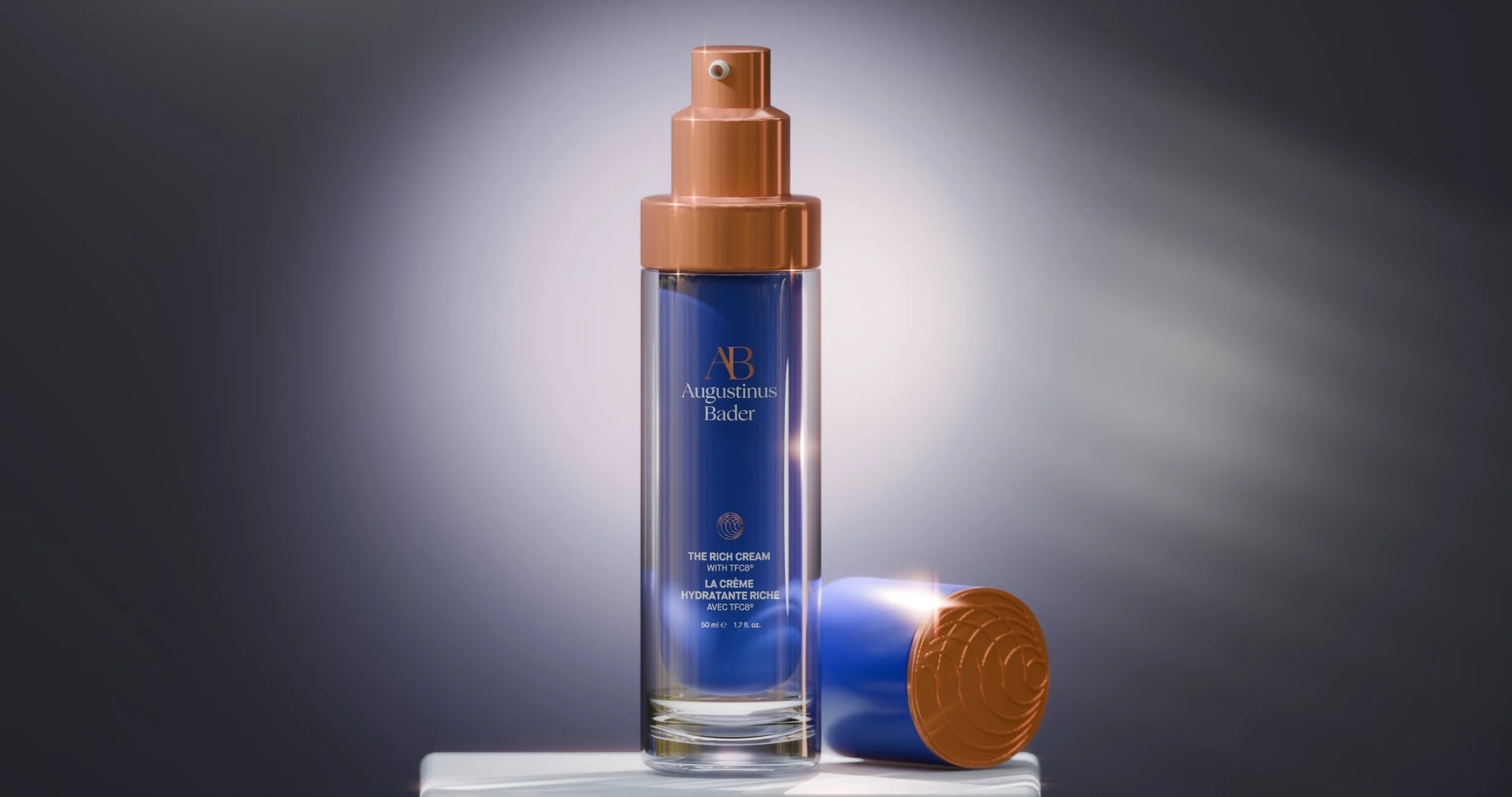 A tall, cylindrical blue bottle with a metallic gold pump dispenser labeled 'Augustinus Bader' and 'The Rich Cream with TFC8' stands on a white platform. A similarly colored cap with an embossed pattern lies next to it. The background is gradient gra