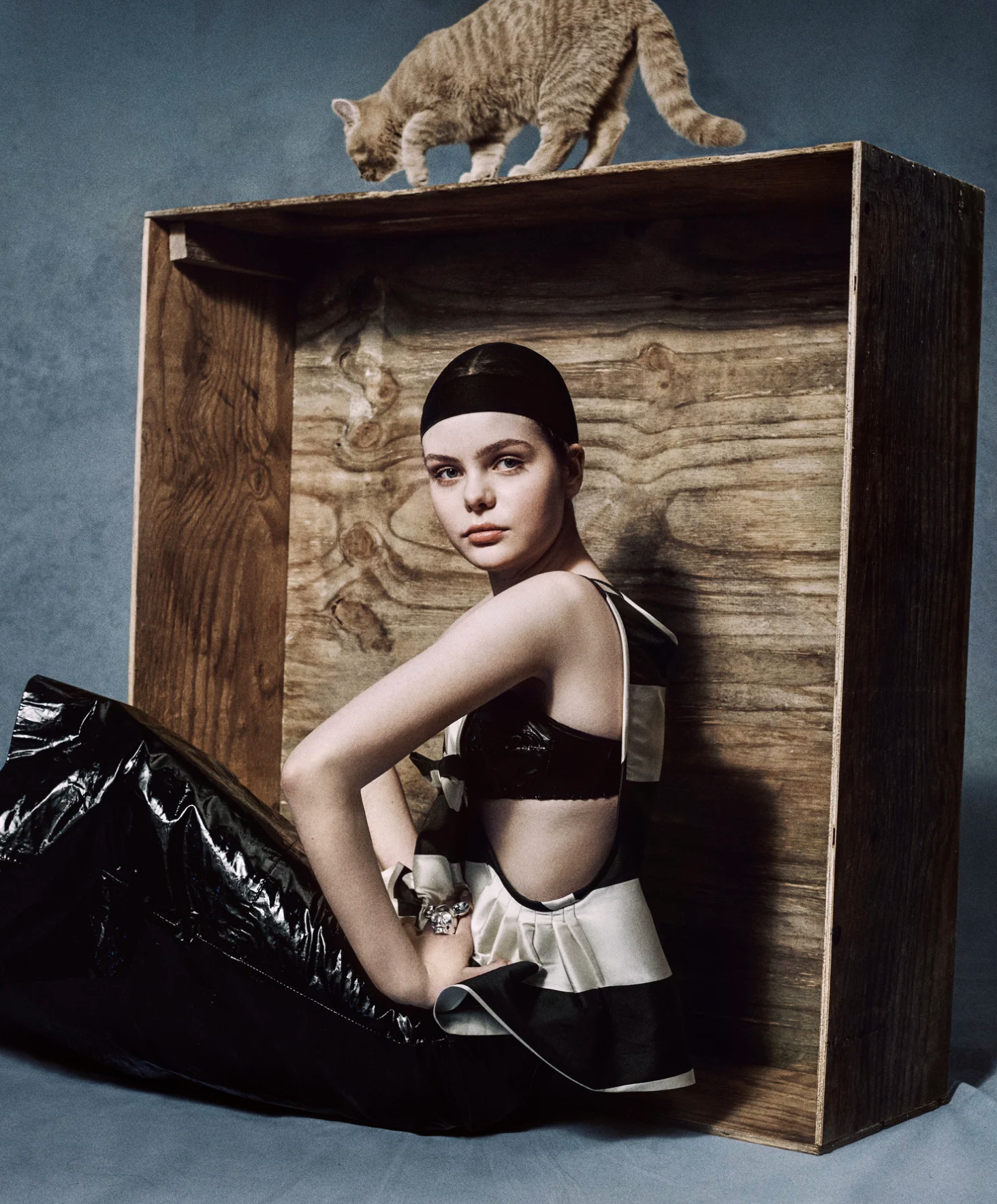 A young woman with dark hair in a sleek updo and light skin, posed sitting in a wooden box with a textured interior. She is wearing a sleeveless black and white dress with bow accents, a wristwatch, and a bracelet, with a neutral expression on her fa