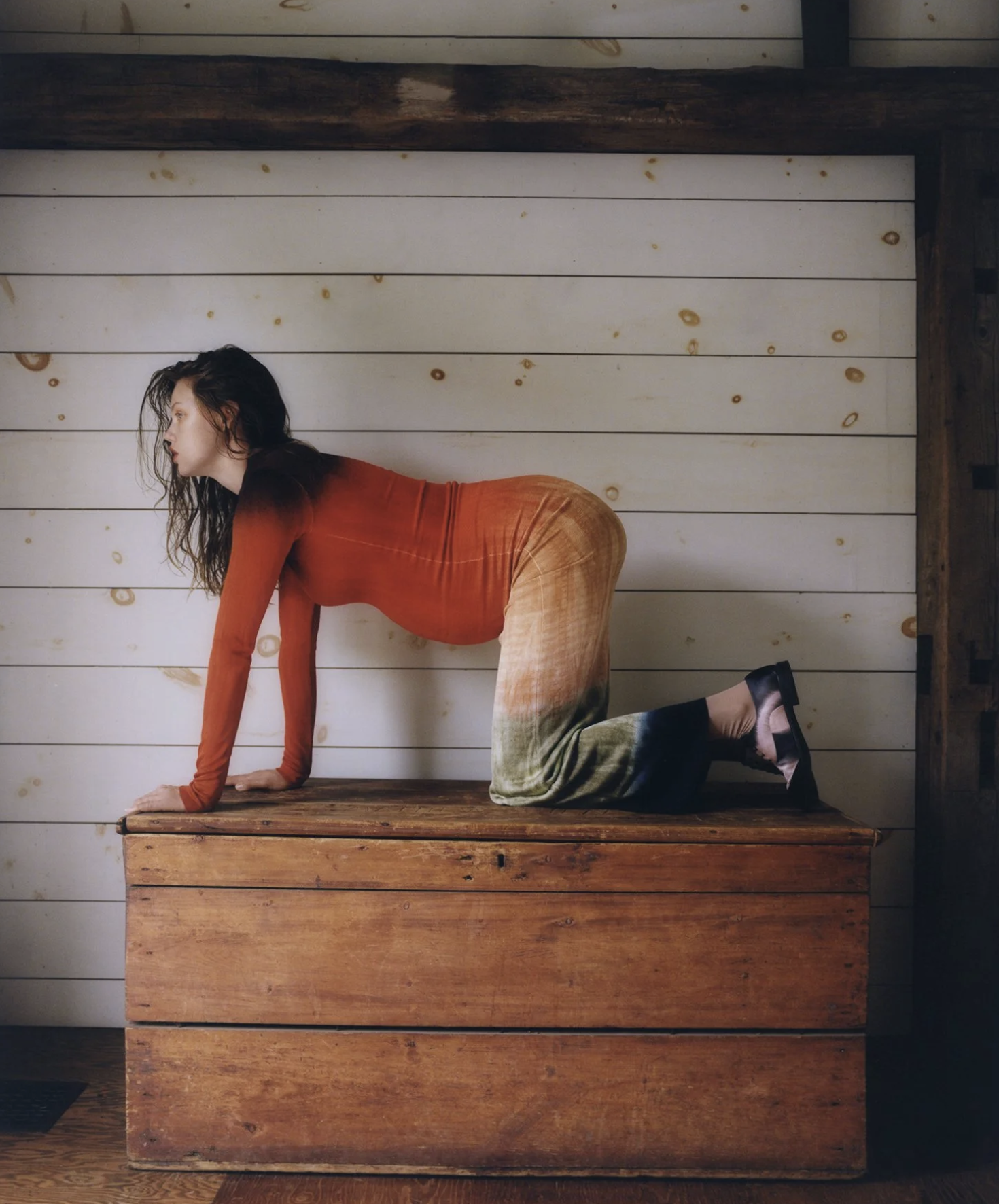 A woman in a red long-sleeve top and patterned skirt crouching on her hands and knees on a wooden surface in a room with white shiplap walls.