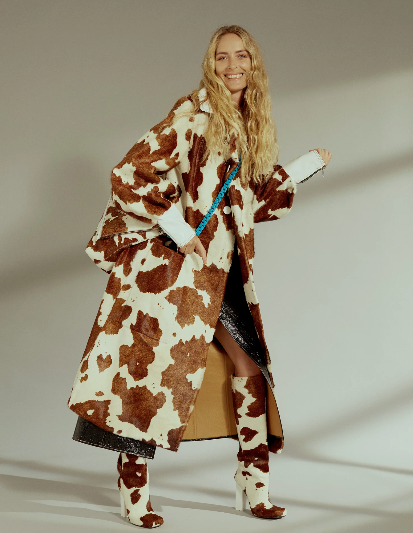 Woman with long blonde curly hair wearing a long cowhide-print coat, matching cowhide-print boots, and black pants with a zipper and a slit, standing against a plain background.