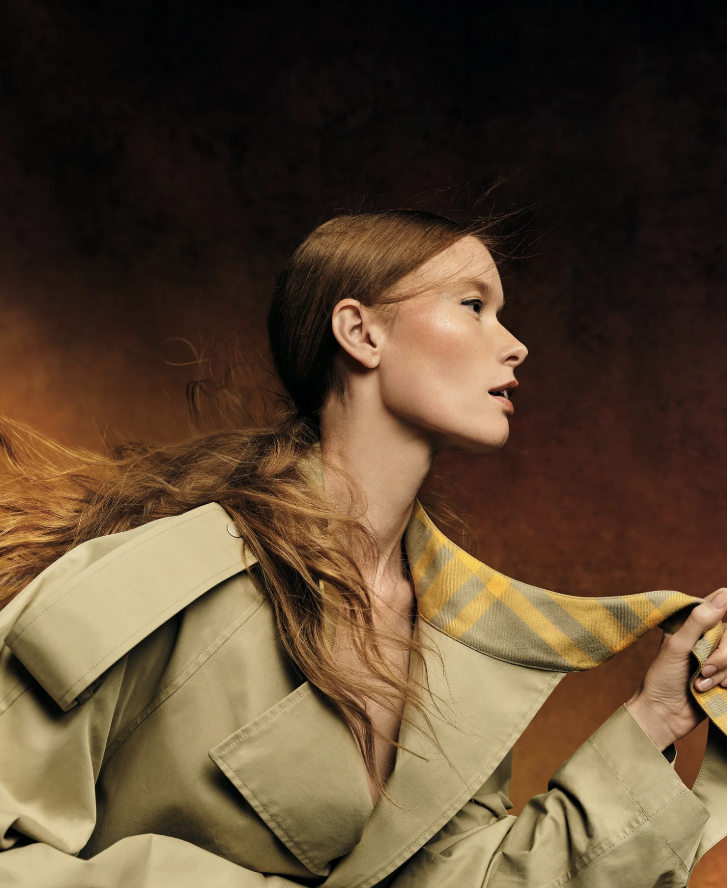 A woman with long, wavy, red hair wearing a beige trench coat with a yellow plaid lining, holding a phone with a colorful background.
