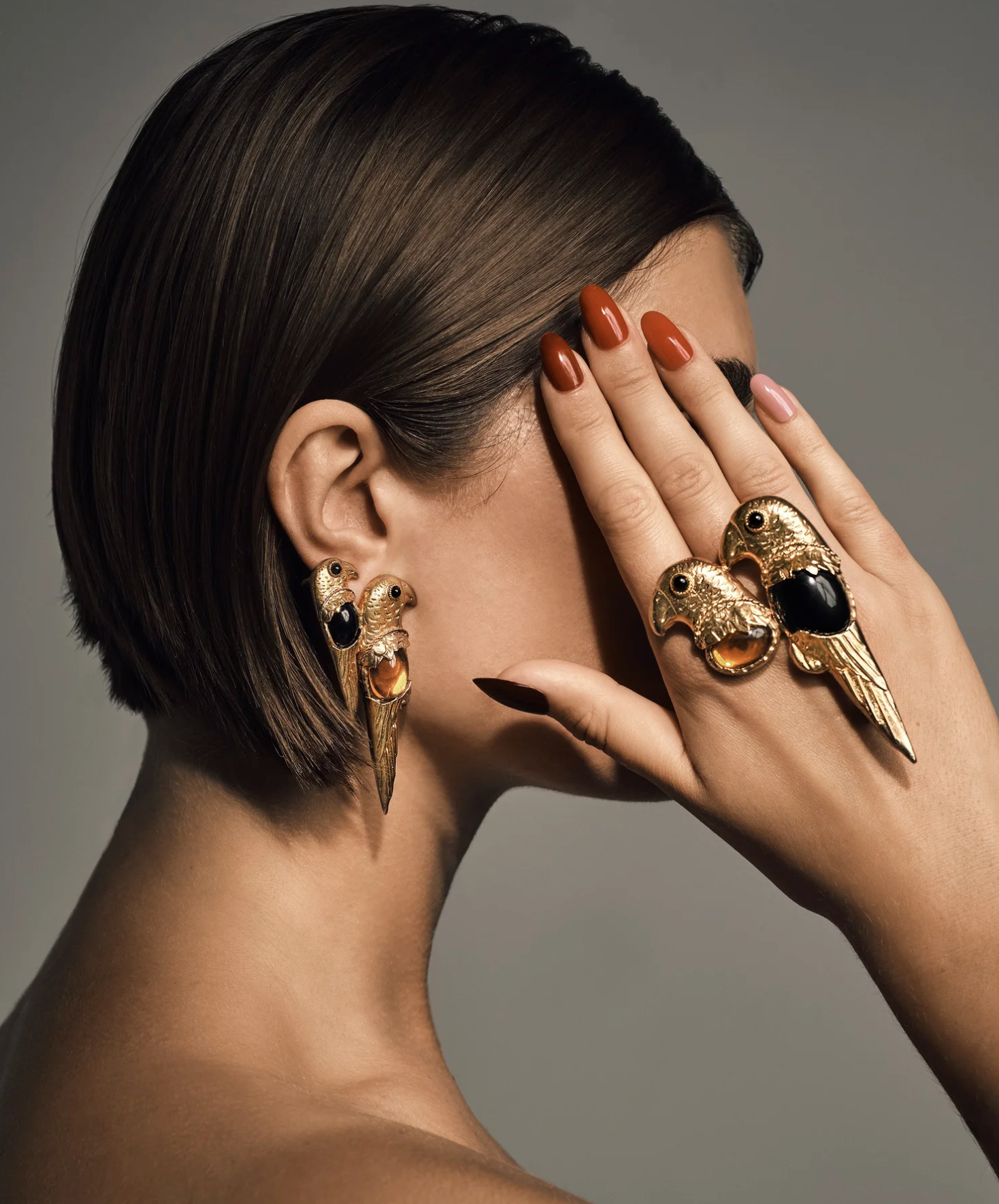 Close-up of a woman with short brown hair, wearing gold earrings and rings. Her hand partially covers her face, showing her manicured nails.