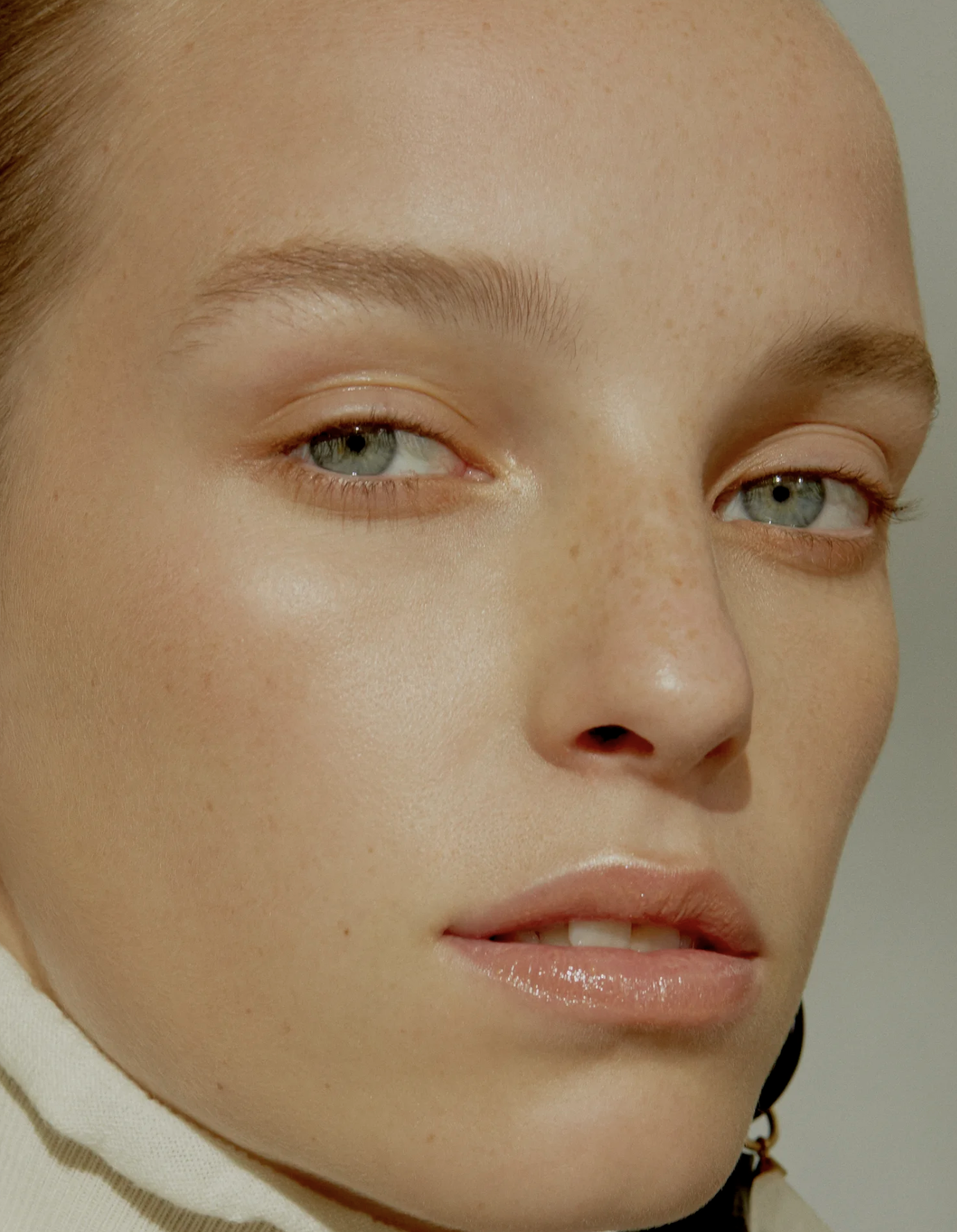 Close-up of a woman's face with blue eyes, light skin, freckles, and minimal makeup, wearing a light-colored top.