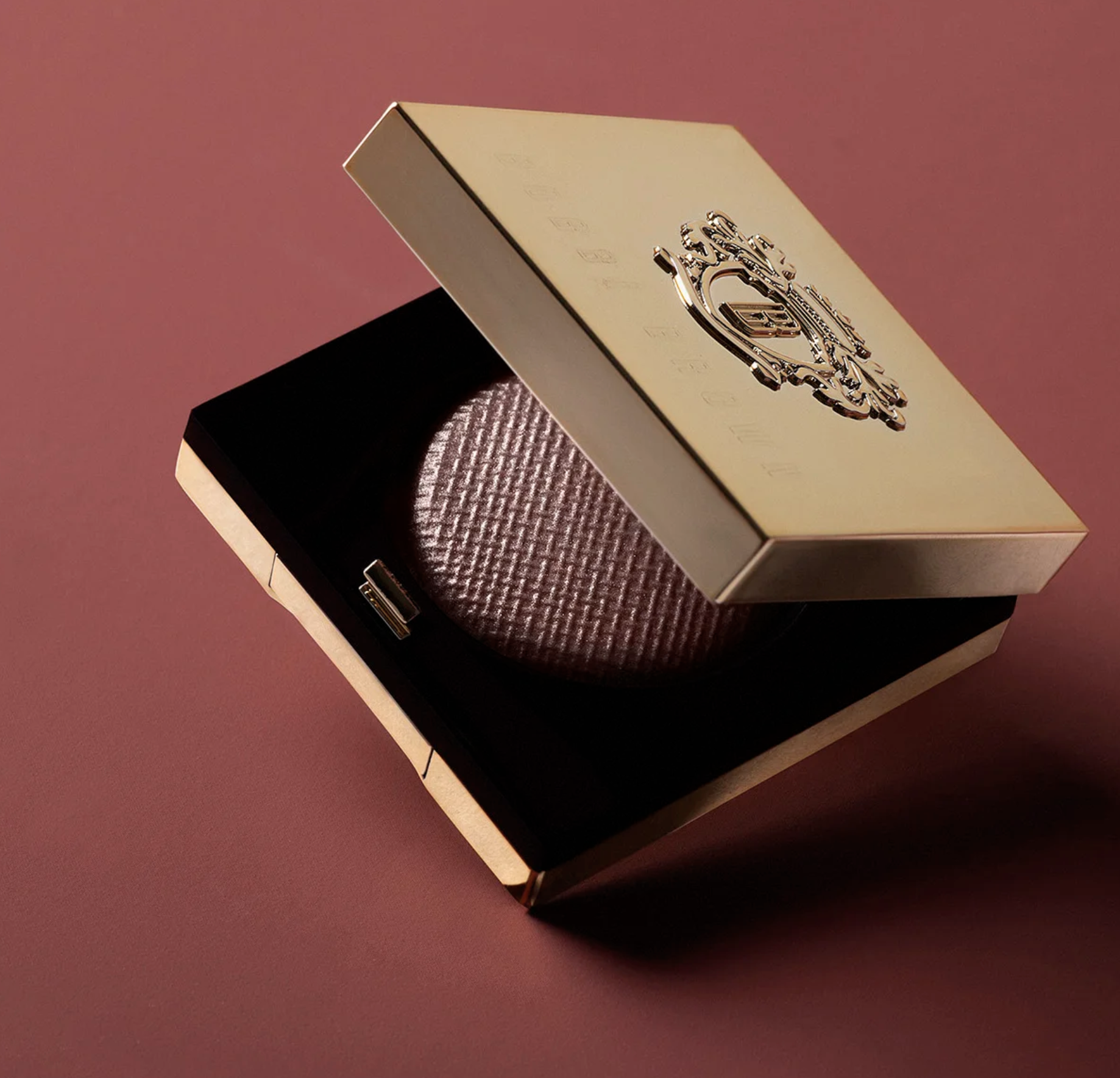 A closed beige box with a metallic emblem on the top, resting on a textured purple and black box, on a pink surface.