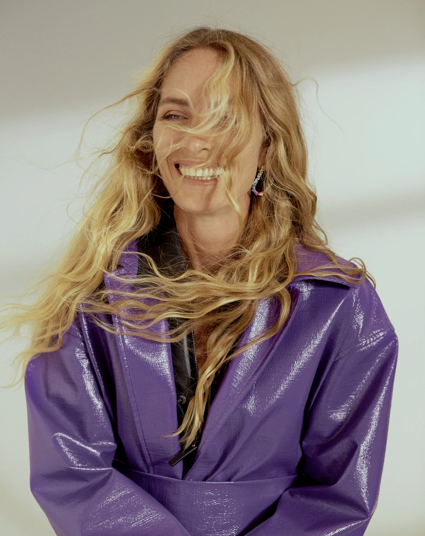 A woman with long wavy blonde hair, smiling and looking to her left, wearing a shiny purple jacket. She has colorful earrings and a black top underneath.