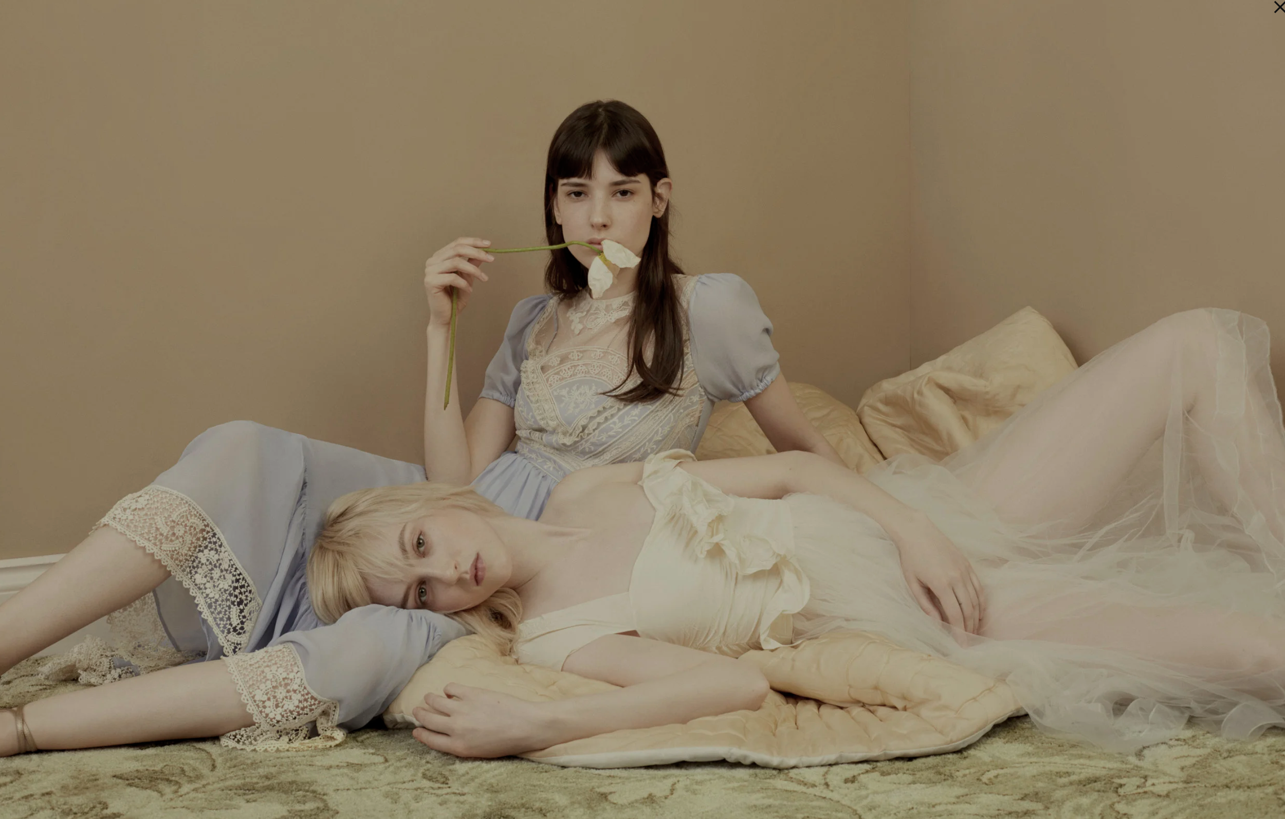 Two women in pastel-colored lingerie, one lying on the bed and the other sitting beside her, with both holding a white flower in their mouths in a softly lit bedroom.