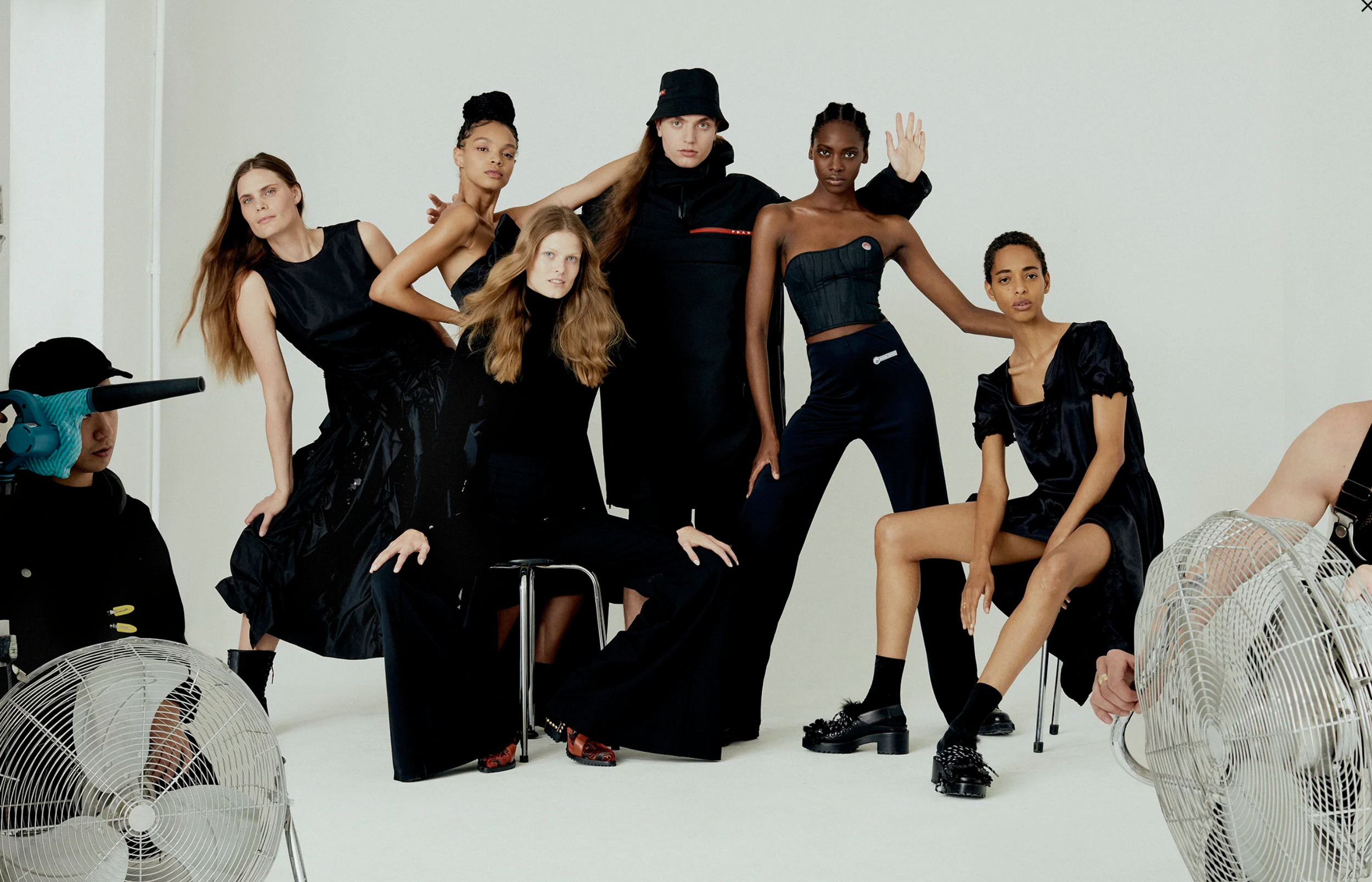 A group of seven diverse female models posing in a studio with a plain white background, some seated and others standing, dressed in black fashion and wearing serious expressions. Three white fans are visible on the floor in the foreground.