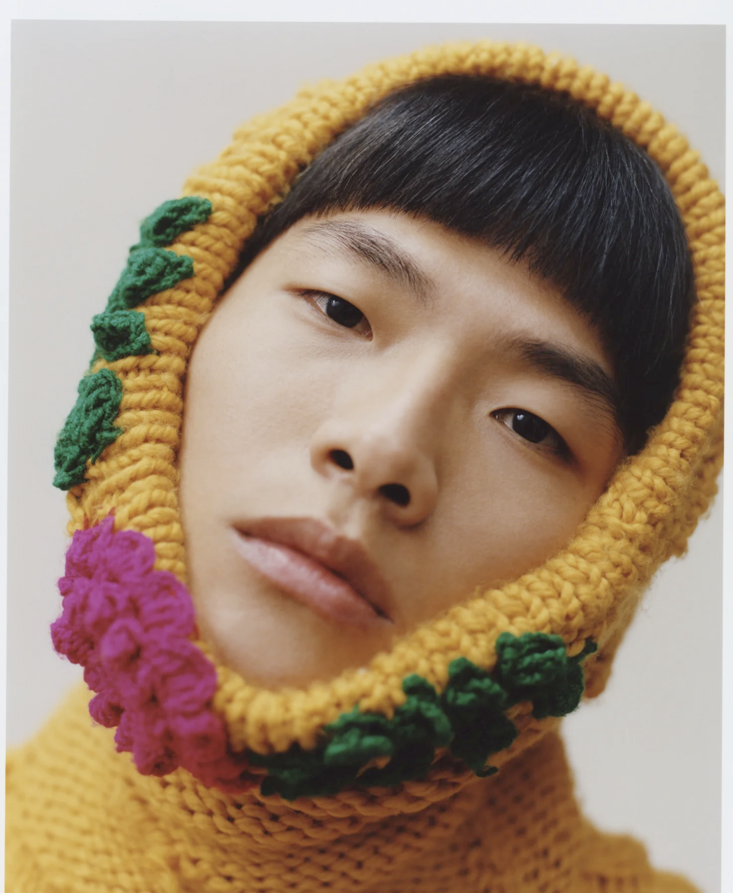Close-up of a person's face with a yellow knit hood decorated with colorful pom-poms, dark hair, and light skin, against a plain background.