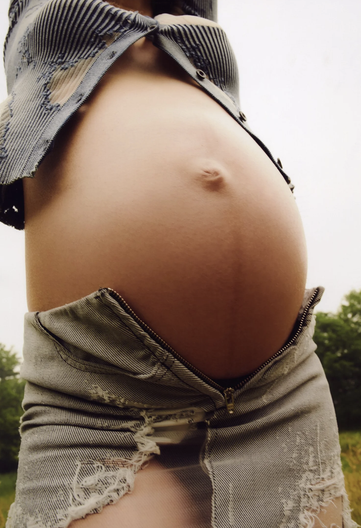 Close-up of a pregnant woman's belly outdoors, with her shirt lifted and shorts unzipped.