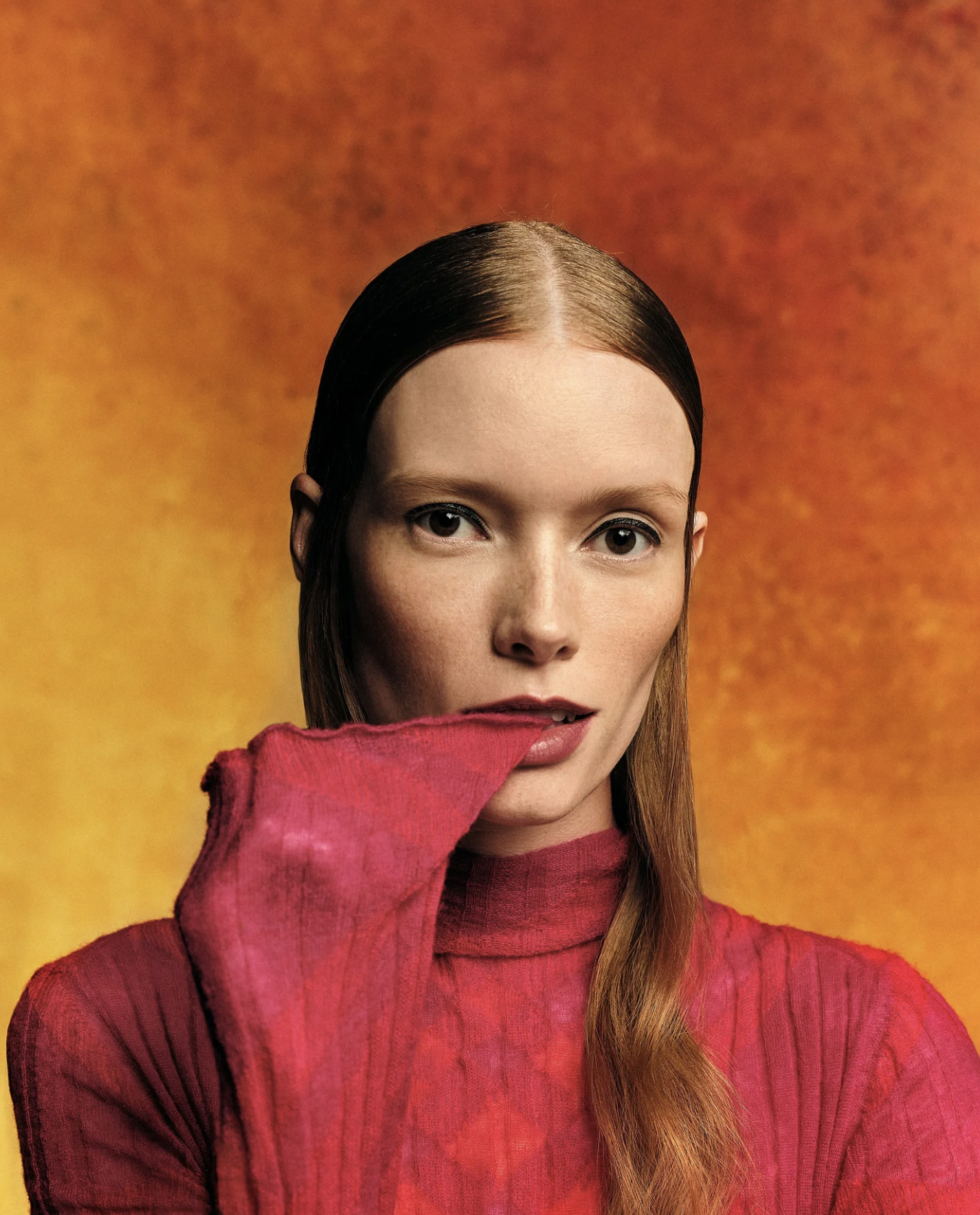A woman with straight brown hair and bright red lipstick wearing a red knit sweater, partially covering her face with her hand, against a gradient orange and yellow background.