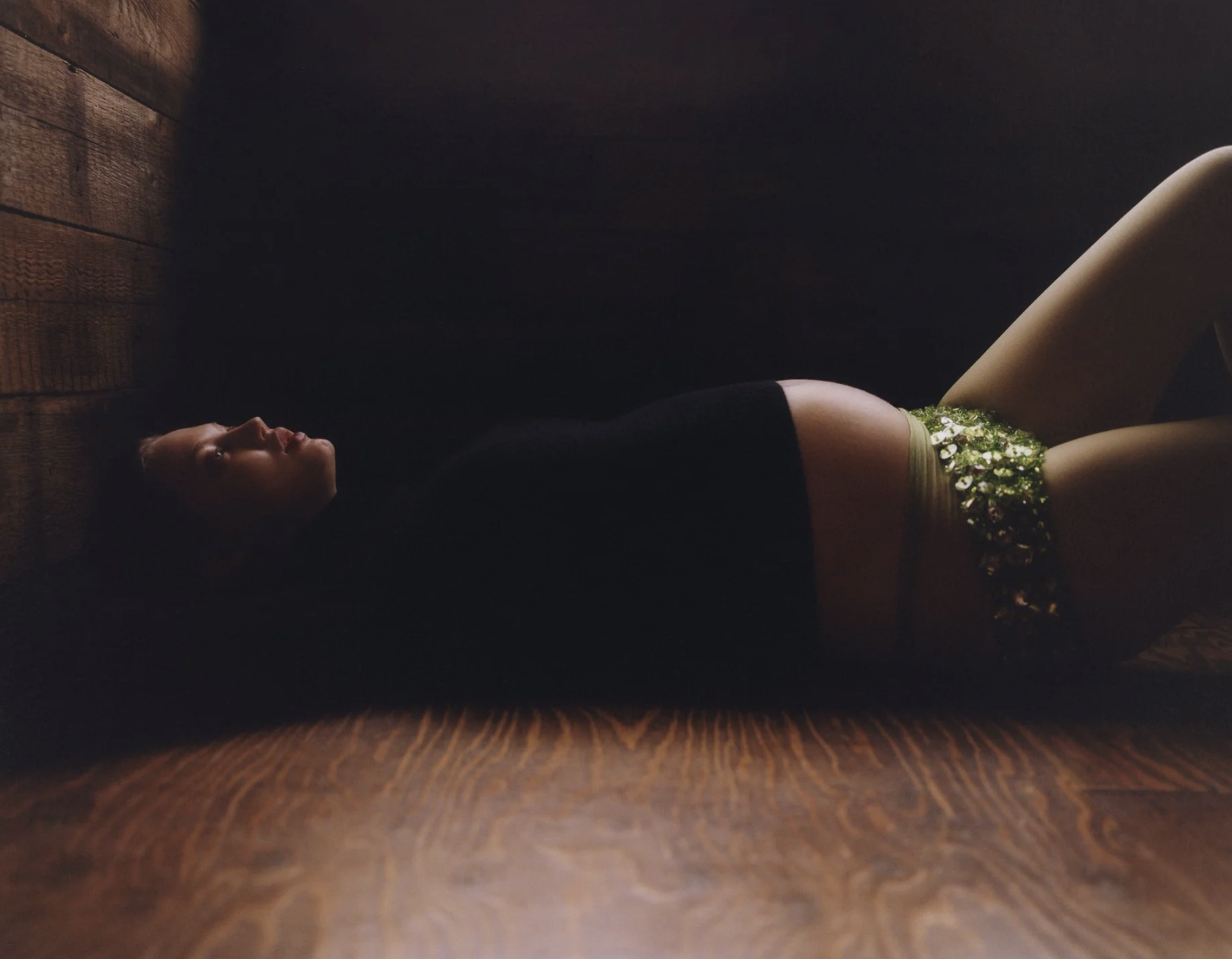 A person lying on their back on a dark floor, illuminated by a ring light, with a wall in the background and a plant near their legs.
