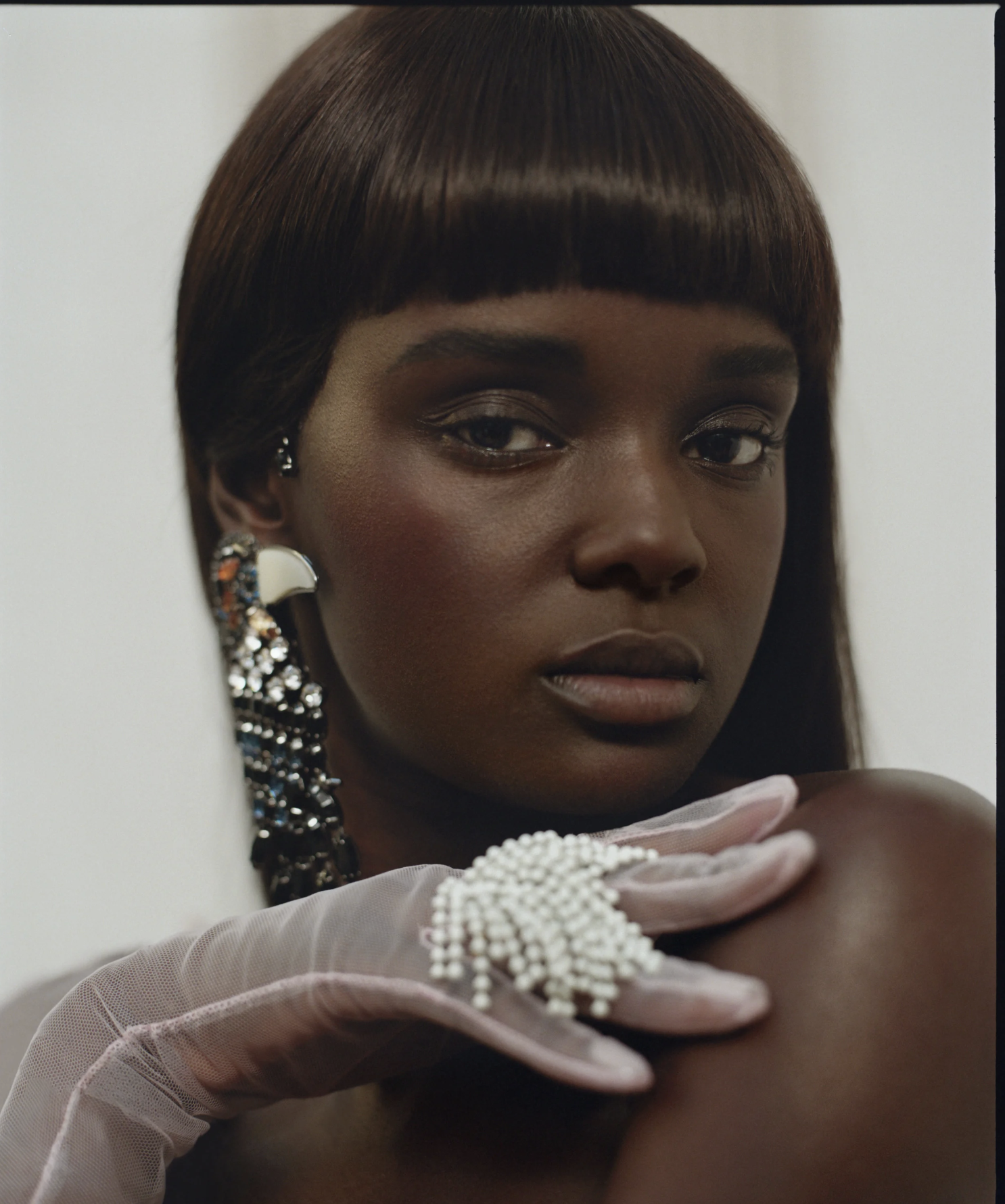A woman with short, straight, dark brown hair and makeup, wearing sparkly earrings, a pearl necklace, and pink gloves, is posing against a plain background.