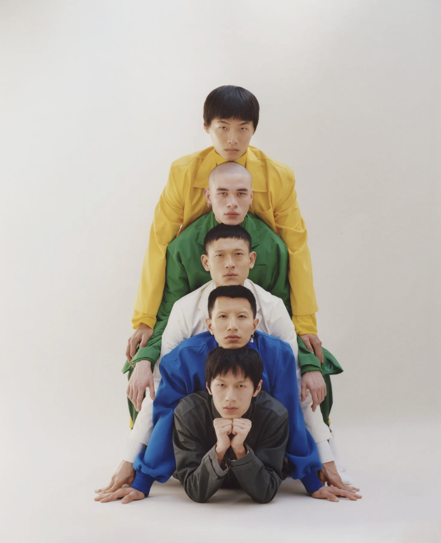 Six young men in colorful jackets arranged in a line, lying on the ground with their hands and feet on the floor, forming a human pyramid against a plain white background.