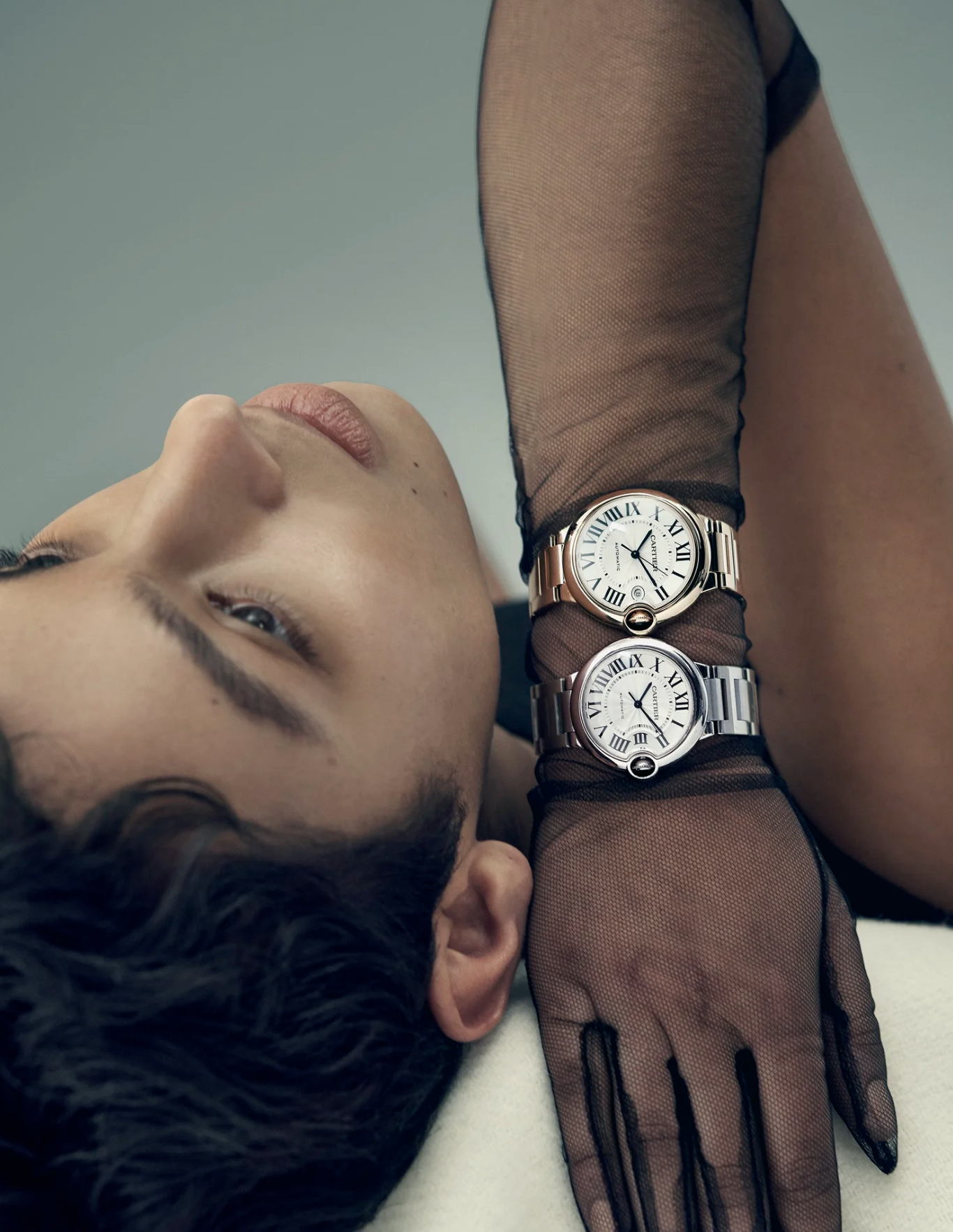 A person with short, dark hair lying on their back, wearing a black sheer top and two silver watches with white faces and Roman numerals on their wrist. The person has a light skin tone and relaxed face.